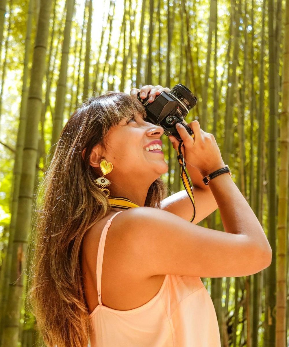 Woman smiles while looking through a camera in a bamboo forest, wearing a light-colored dress and earrings.
