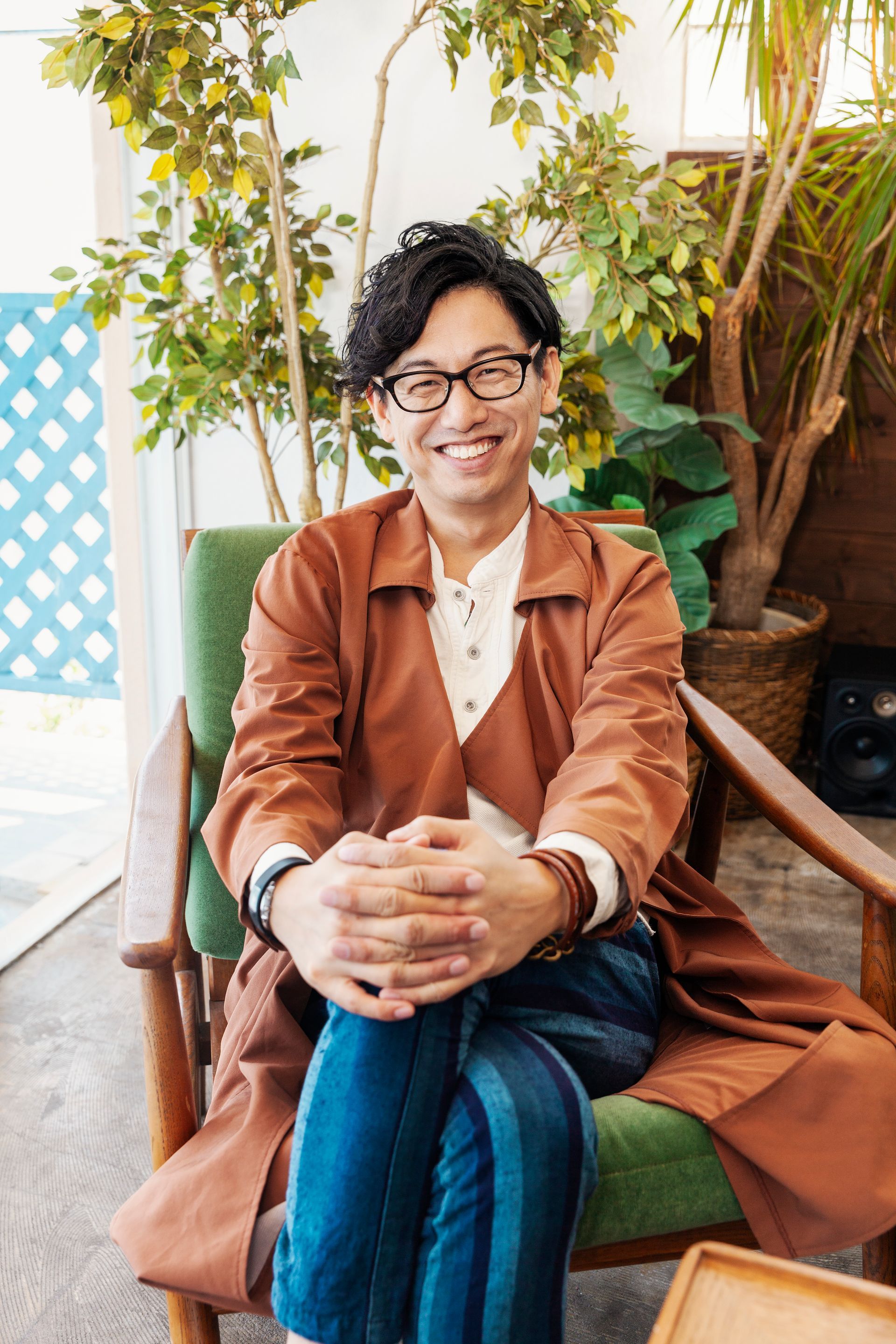 Man in glasses smiles, sitting in green chair, wearing brown jacket and blue pants, with plants in background.