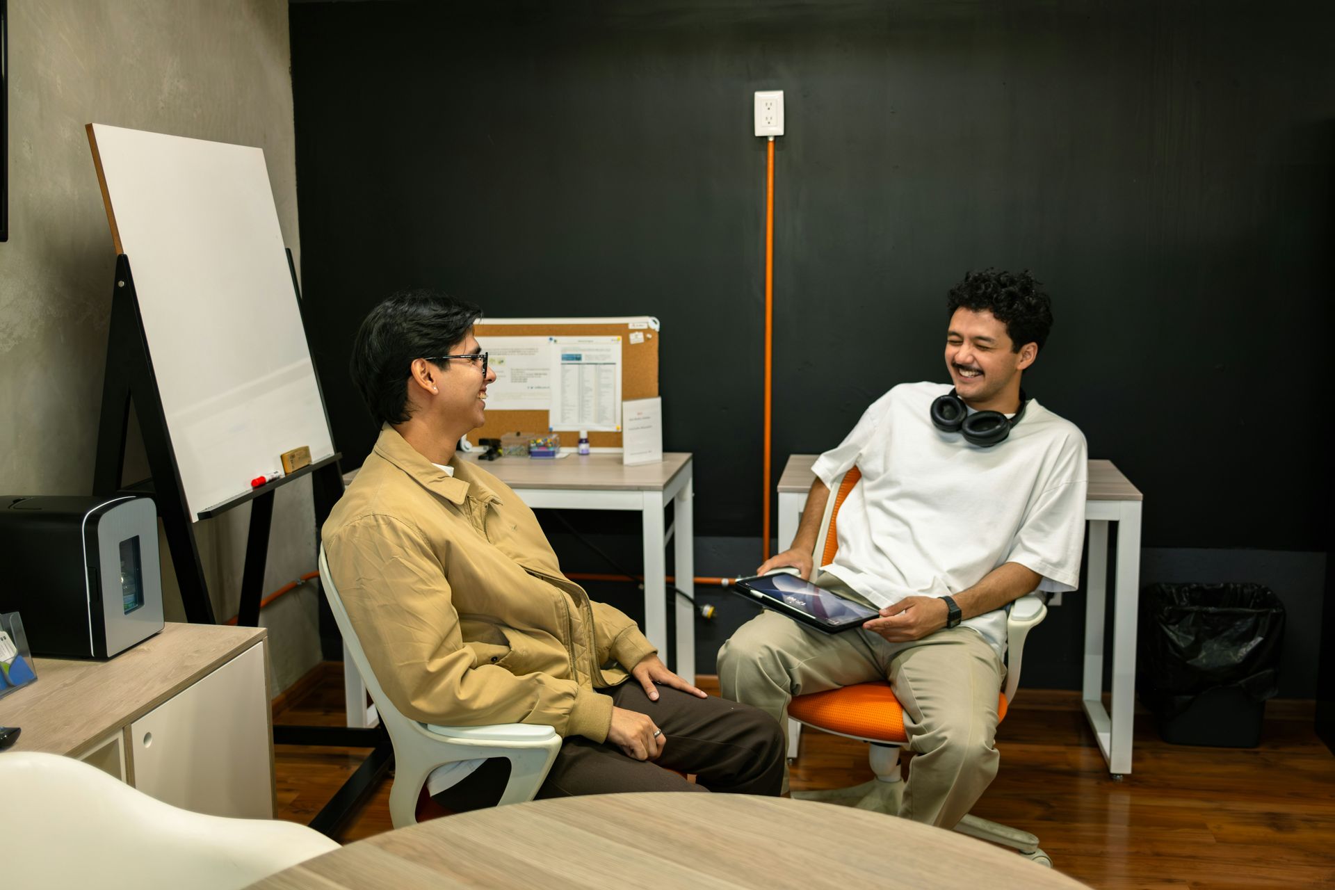 Two men in an office, one holding a tablet and smiling, the other looking at him, both seated.