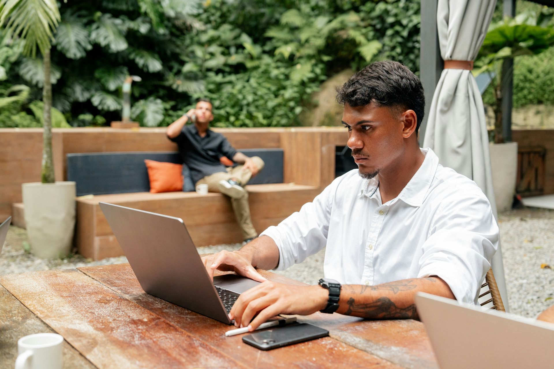 Man working on laptop at outdoor table; another person relaxes in the background.