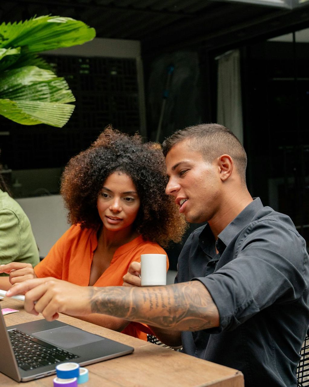 Man points to a laptop screen, explaining something to a woman holding a mug. They sit at a table outdoors.