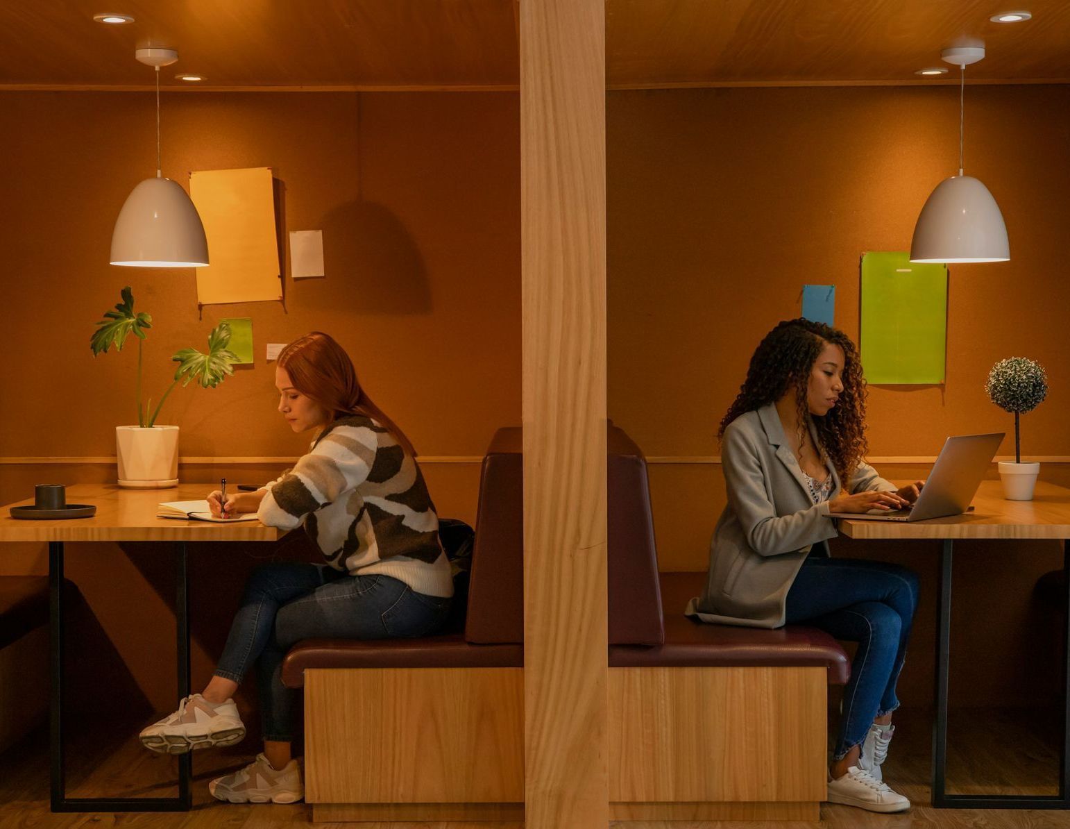 Two women work in separate office booths, one writing, the other typing on a laptop.