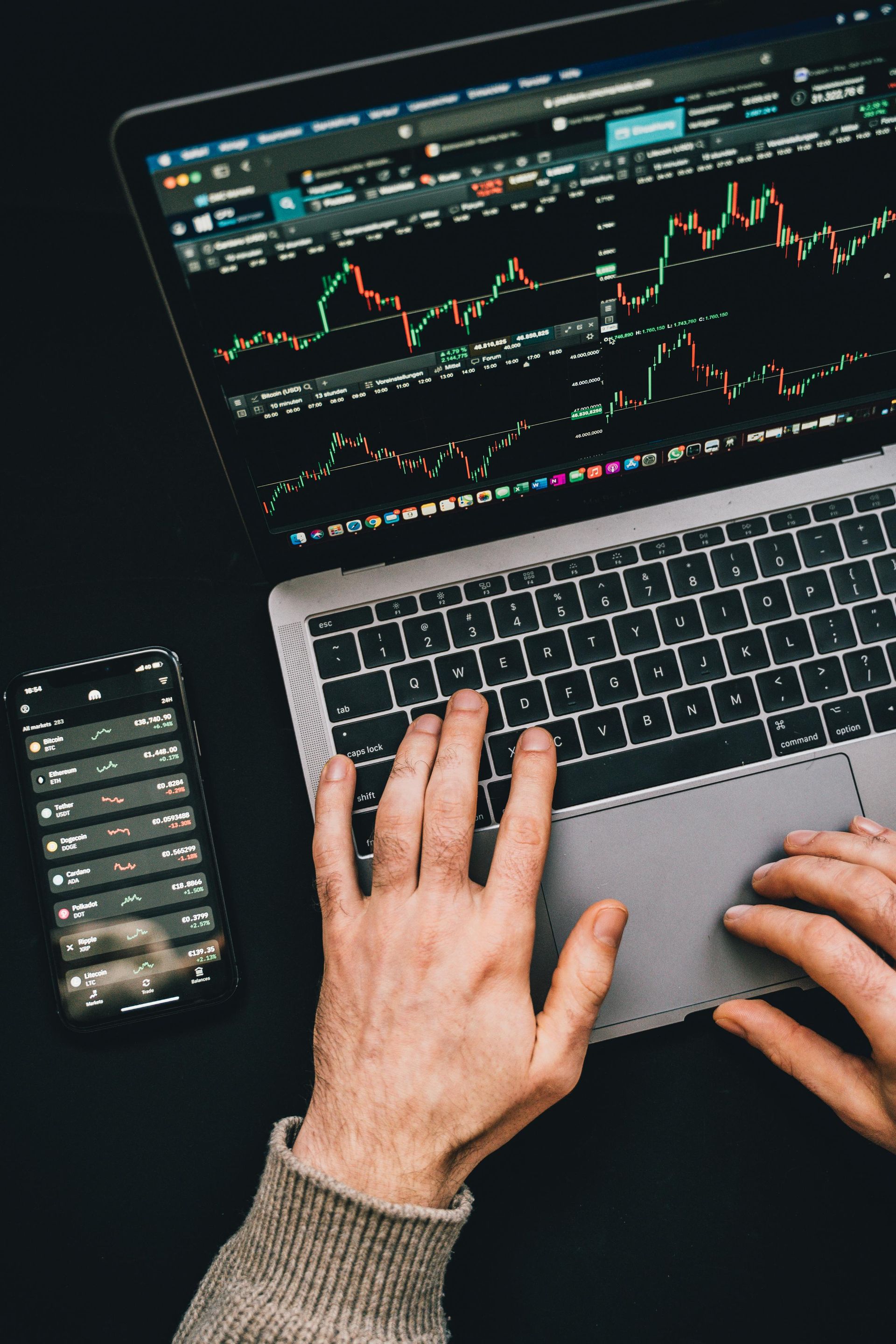 Hands on laptop with stock charts; phone displays stock data on black surface.