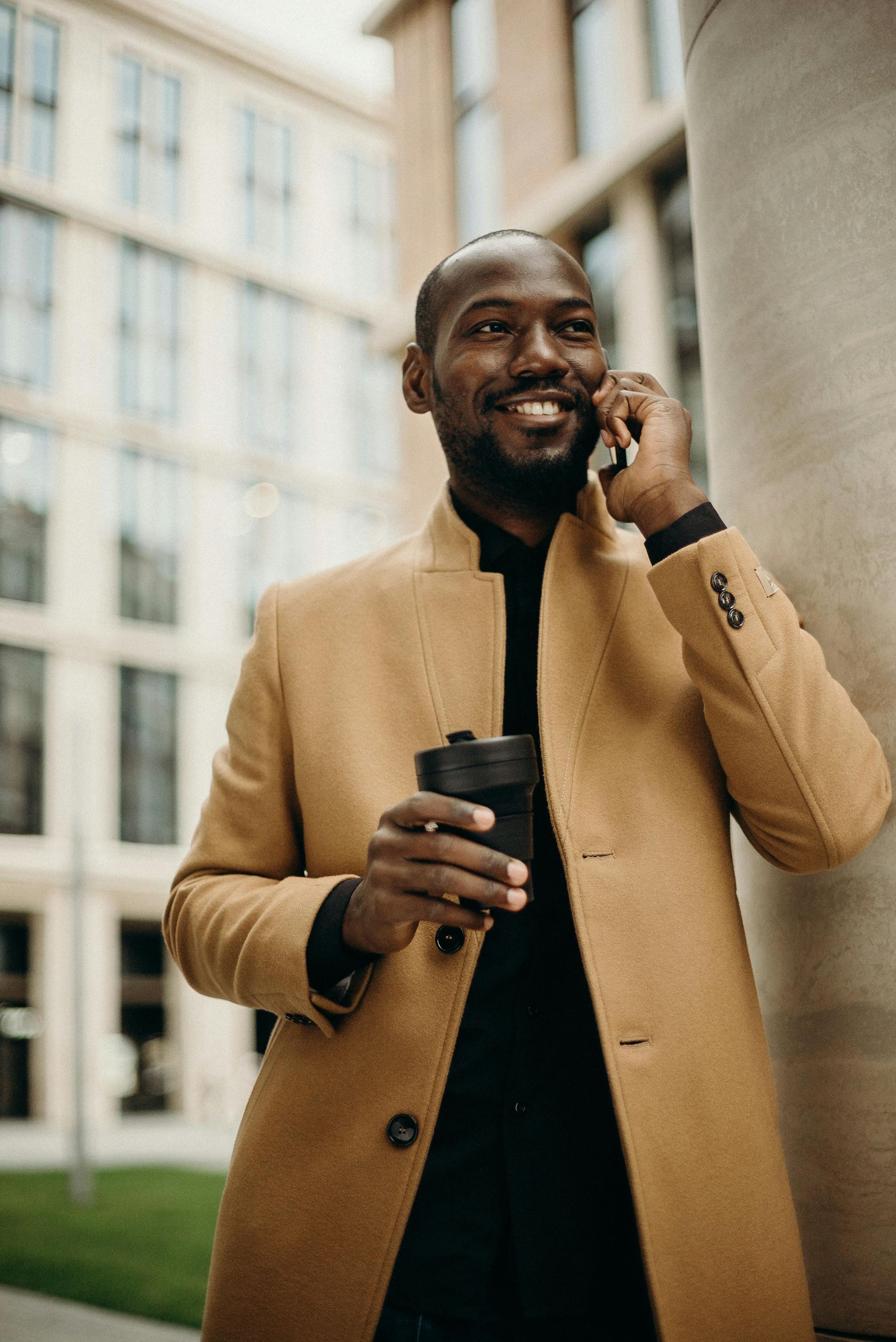 Man in tan coat smiles while on phone, holding coffee near a building.