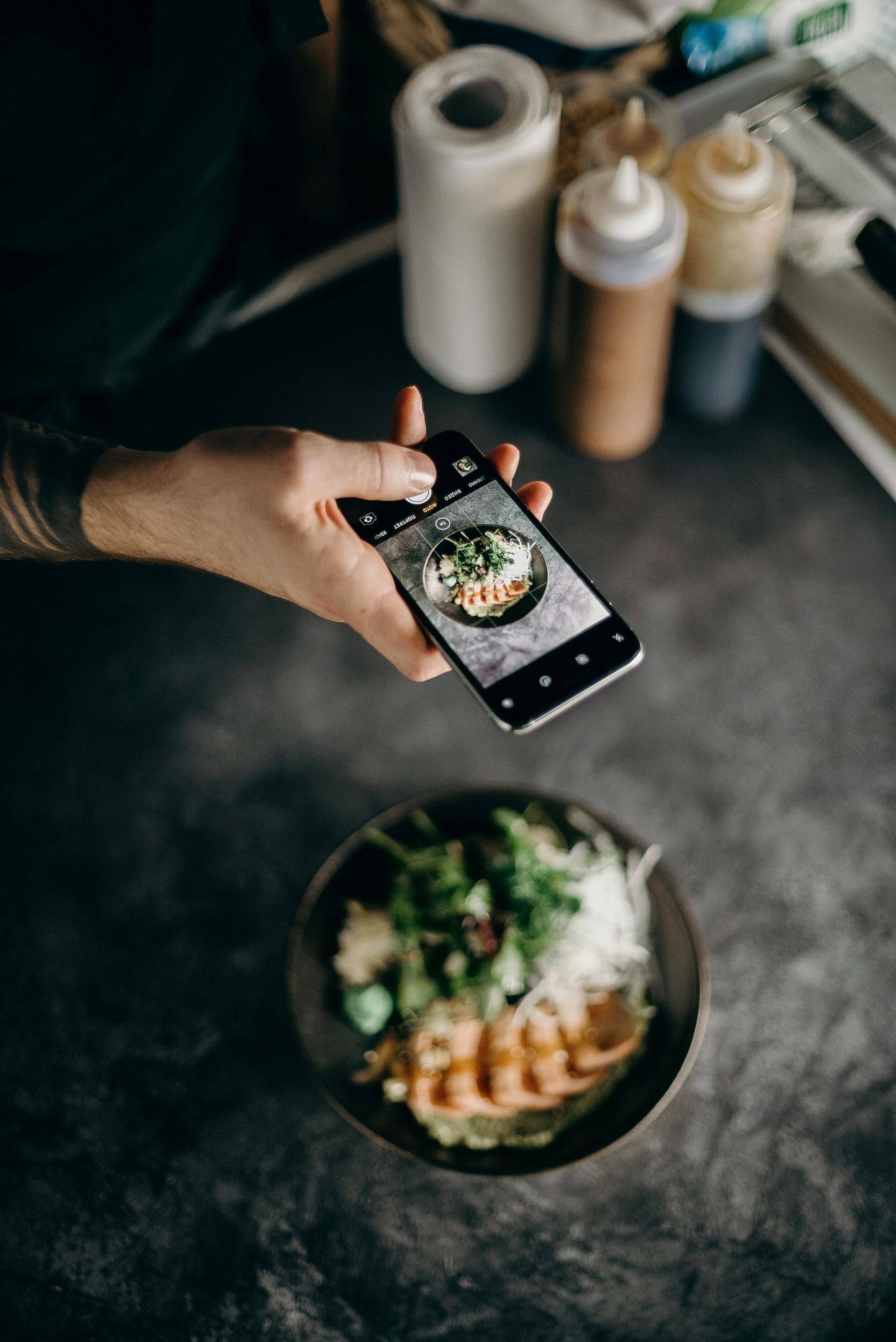 A hand holding a phone, taking a photo of a plate of food. The plate is on a dark surface.