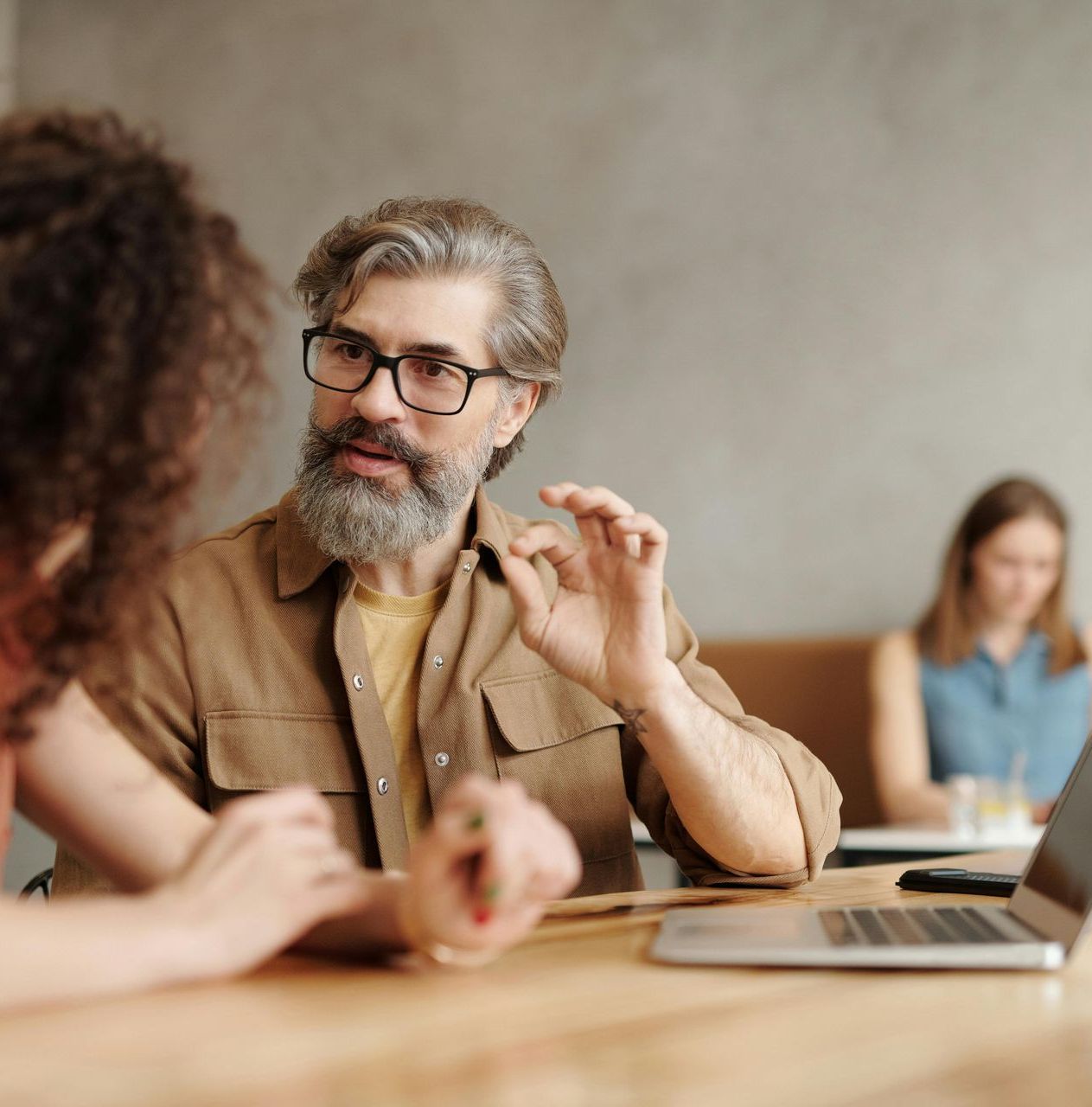 Man gesturing while speaking to a person at a table with a laptop, another person in the background.