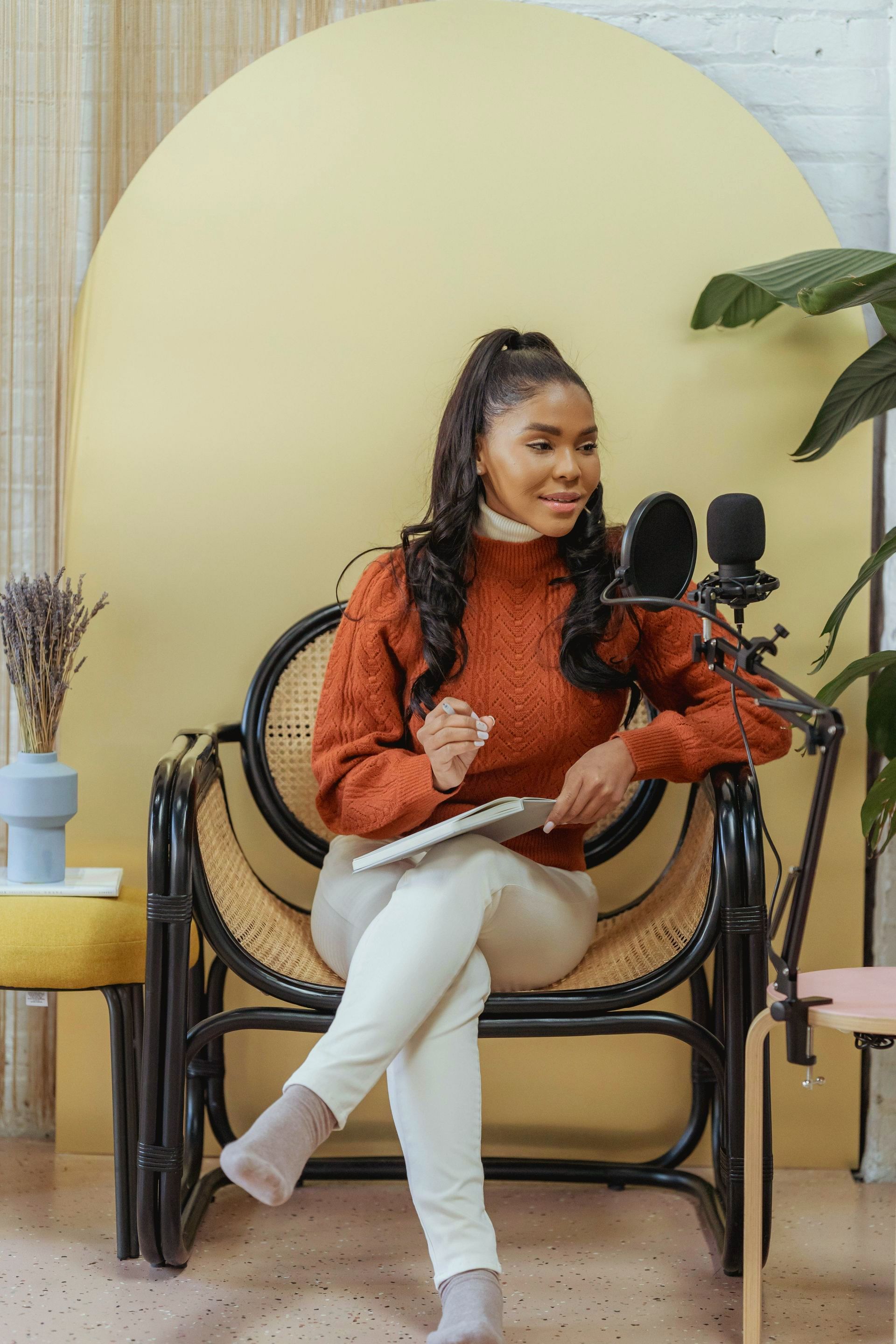 Woman in rust-colored sweater sits in chair, speaking into microphone. Yellow background, holding a tablet.