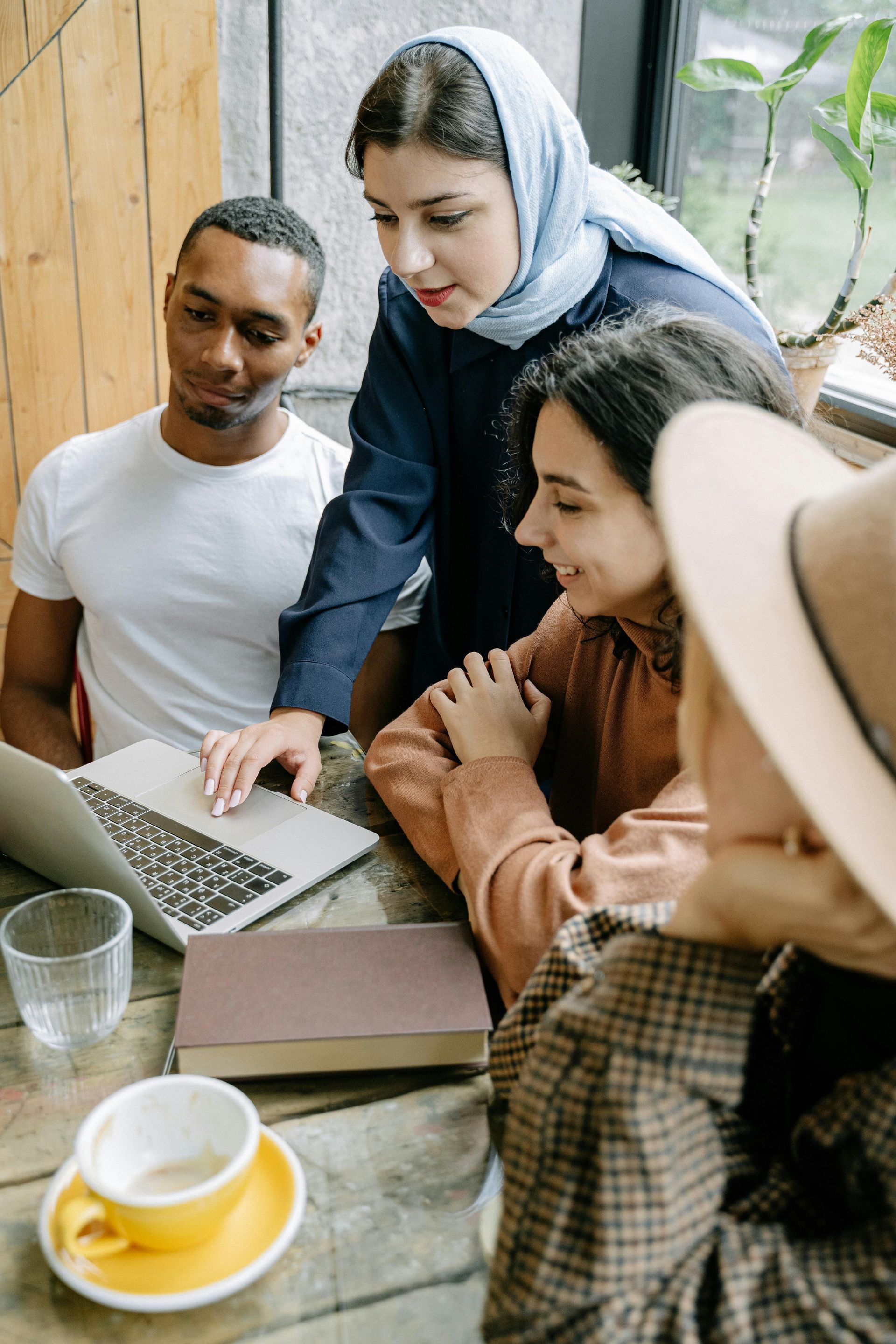 Four people around a table looking at a laptop. Woman in blue headscarf leans in, others smile. Books and coffee are present.