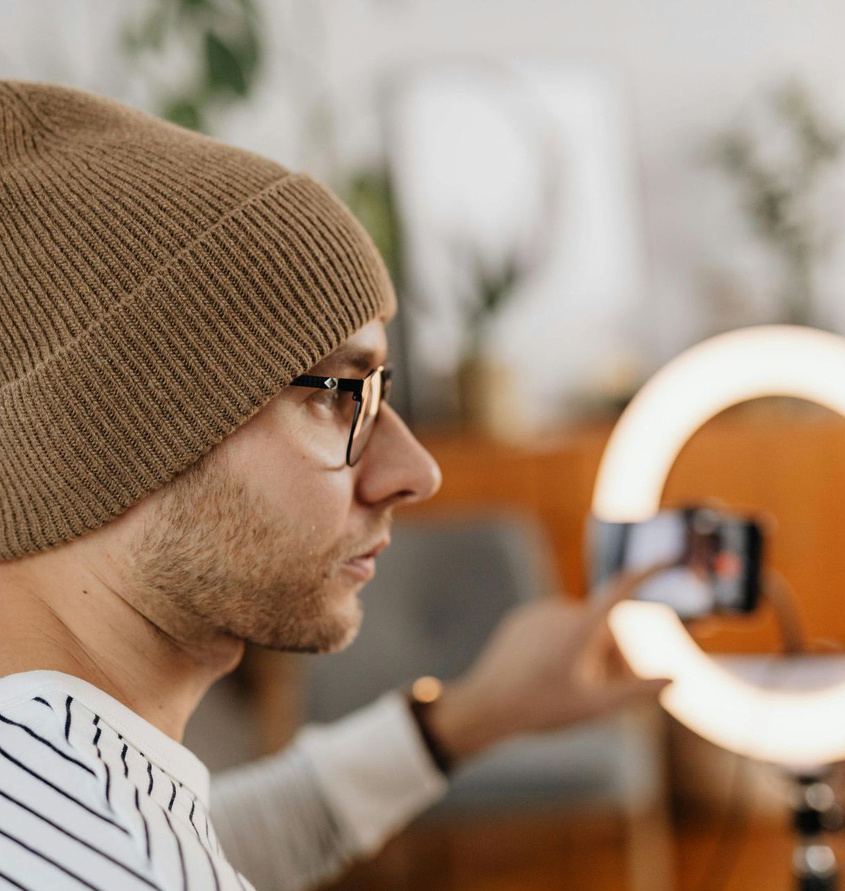 Man wearing glasses and a beanie films with a phone and ring light indoors.