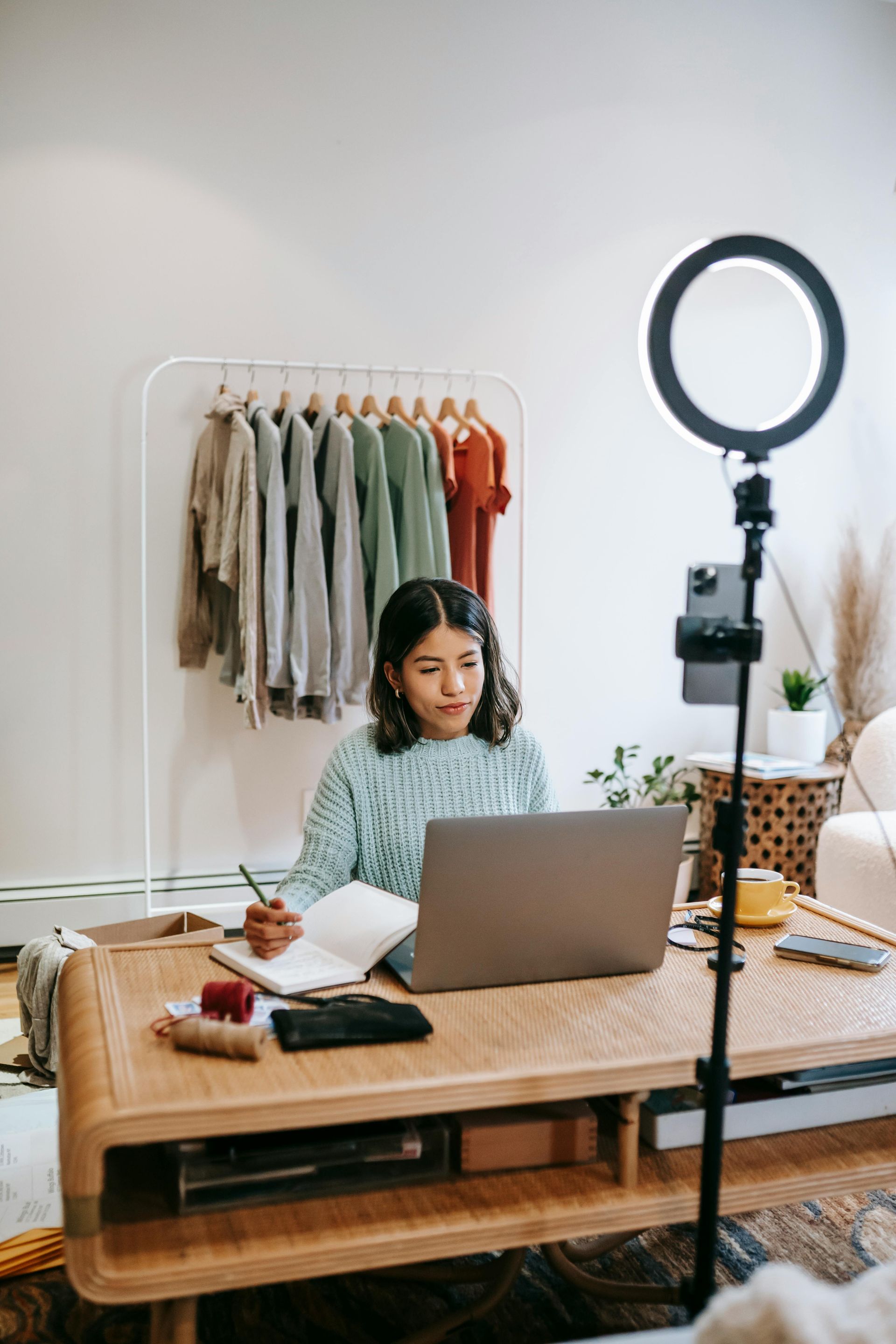 Woman at desk, working on laptop, with ring light and clothes rack in background.