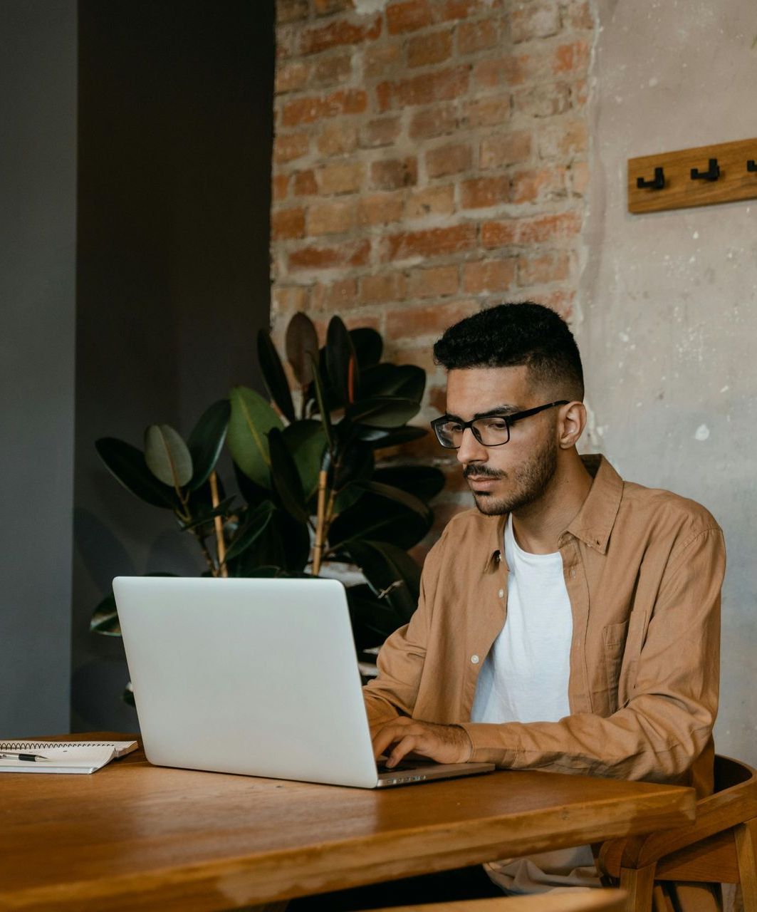 Man at wooden table working on laptop in front of brick wall, plant in background.