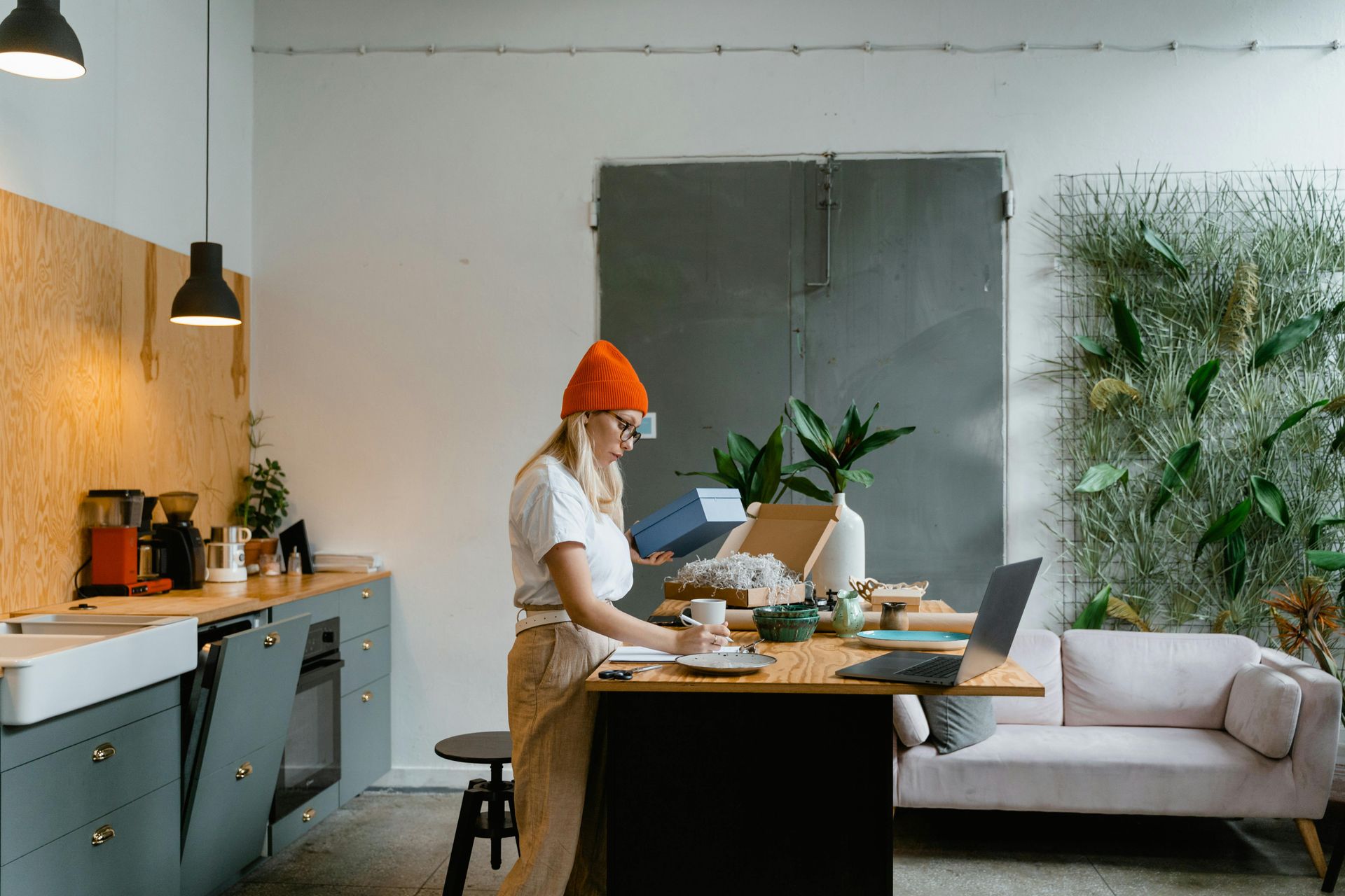 Woman with orange hat working at a desk in a modern kitchen. She is opening a box near a laptop and plants.