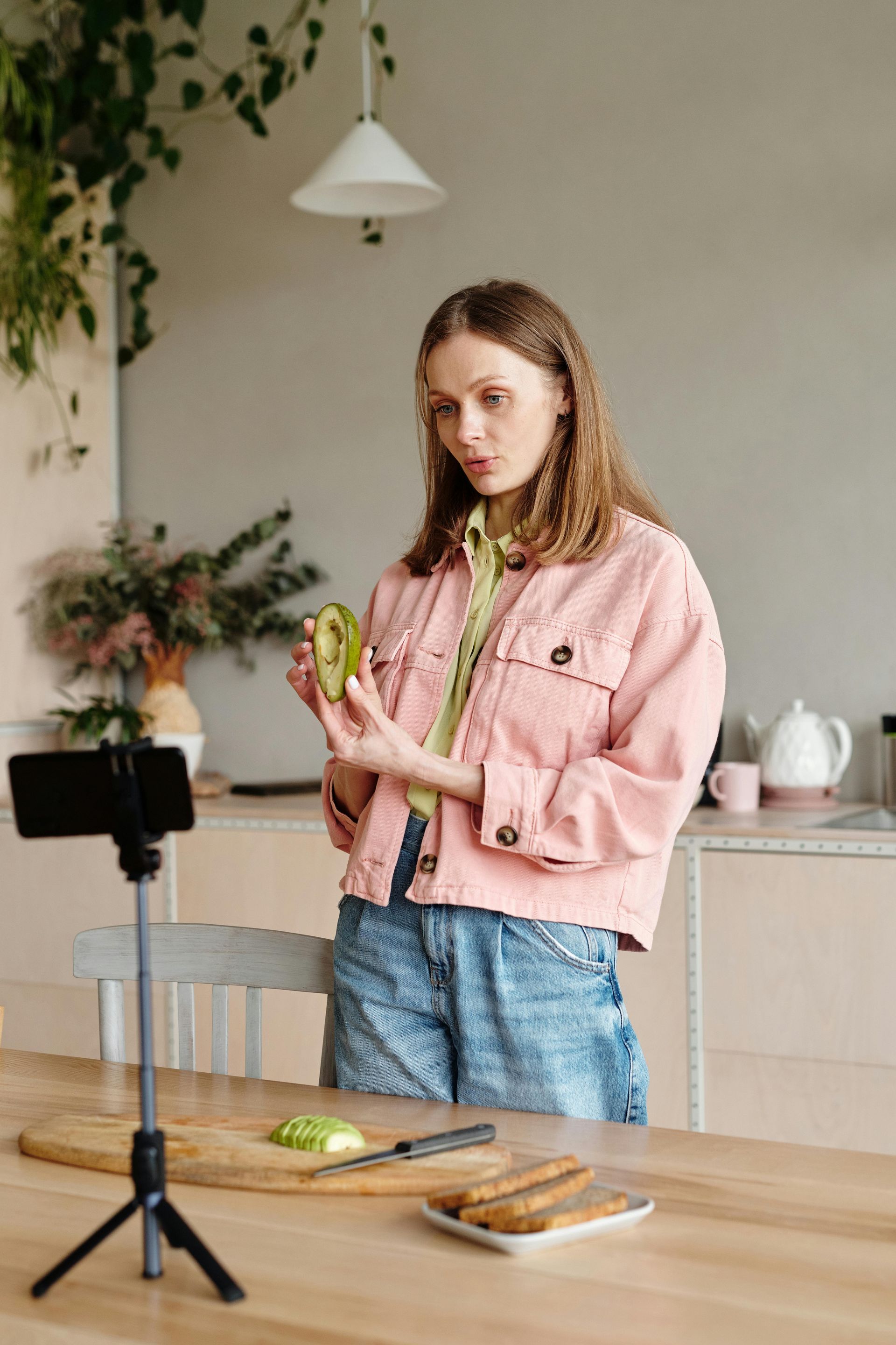 Woman in pink jacket, holding kiwi, filming herself in kitchen.