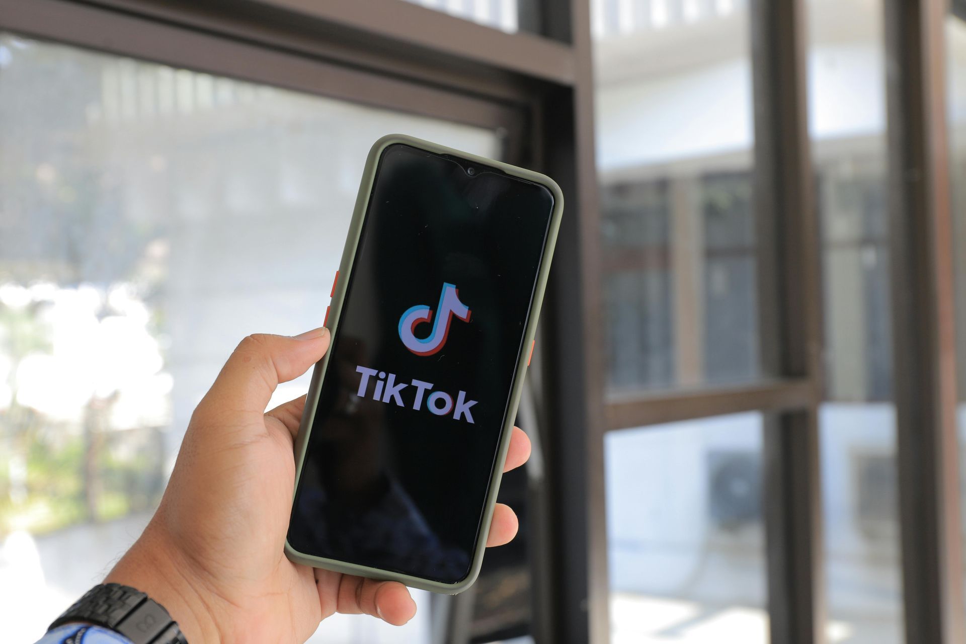Hand holding a smartphone displaying the TikTok logo. Green phone case, window in the background.