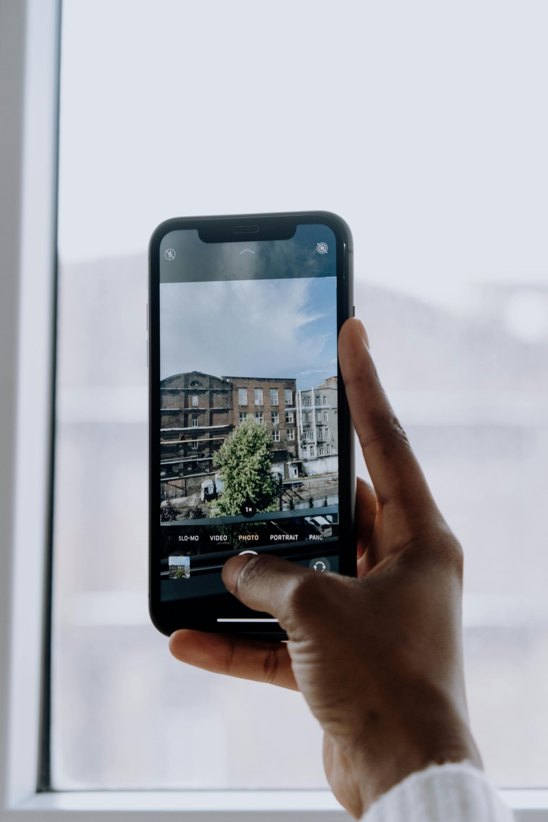 A hand holding a phone, taking a photo of buildings and a tree outside a window.