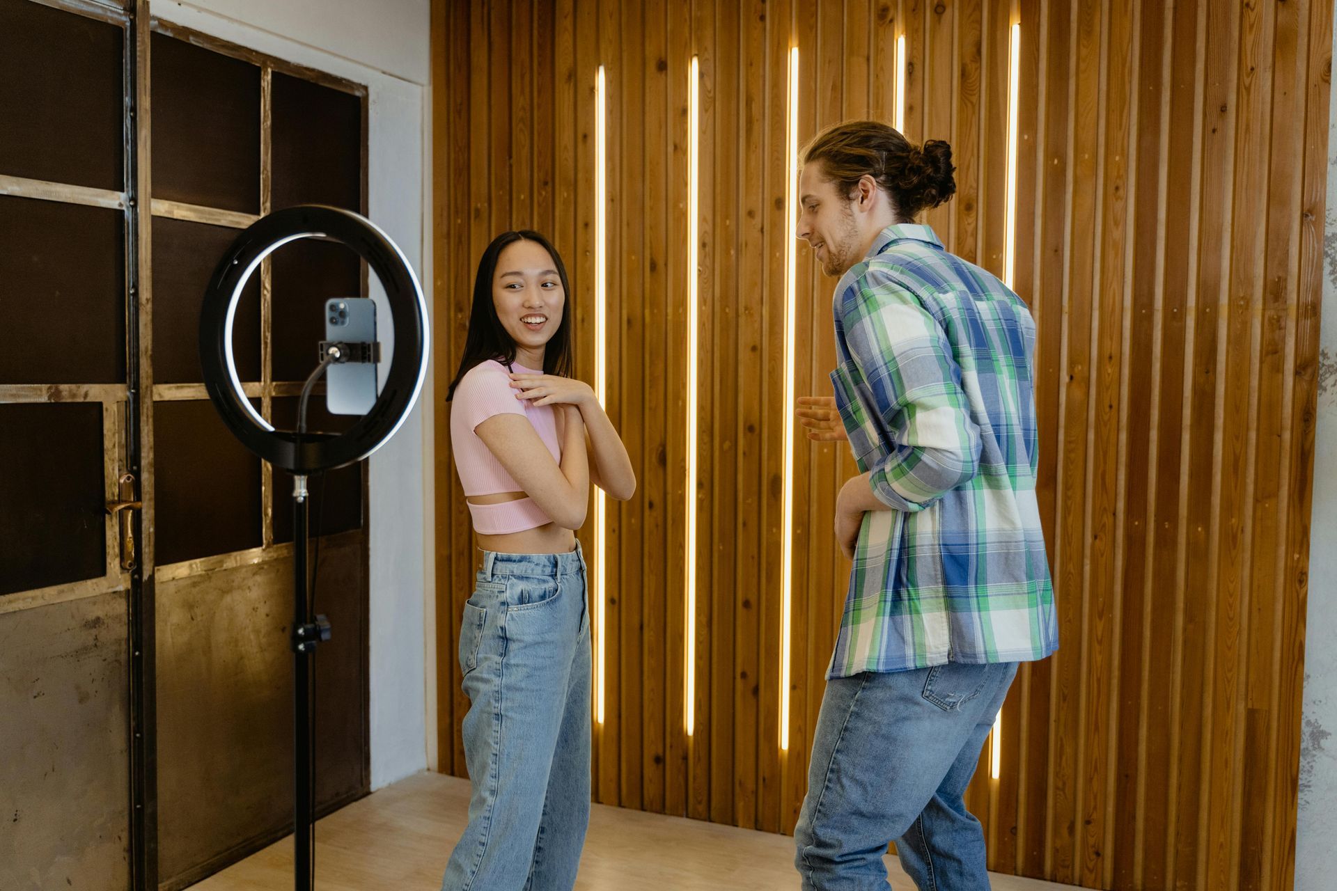 Woman and man dancing near a ring light and phone; wood background with vertical lights.
