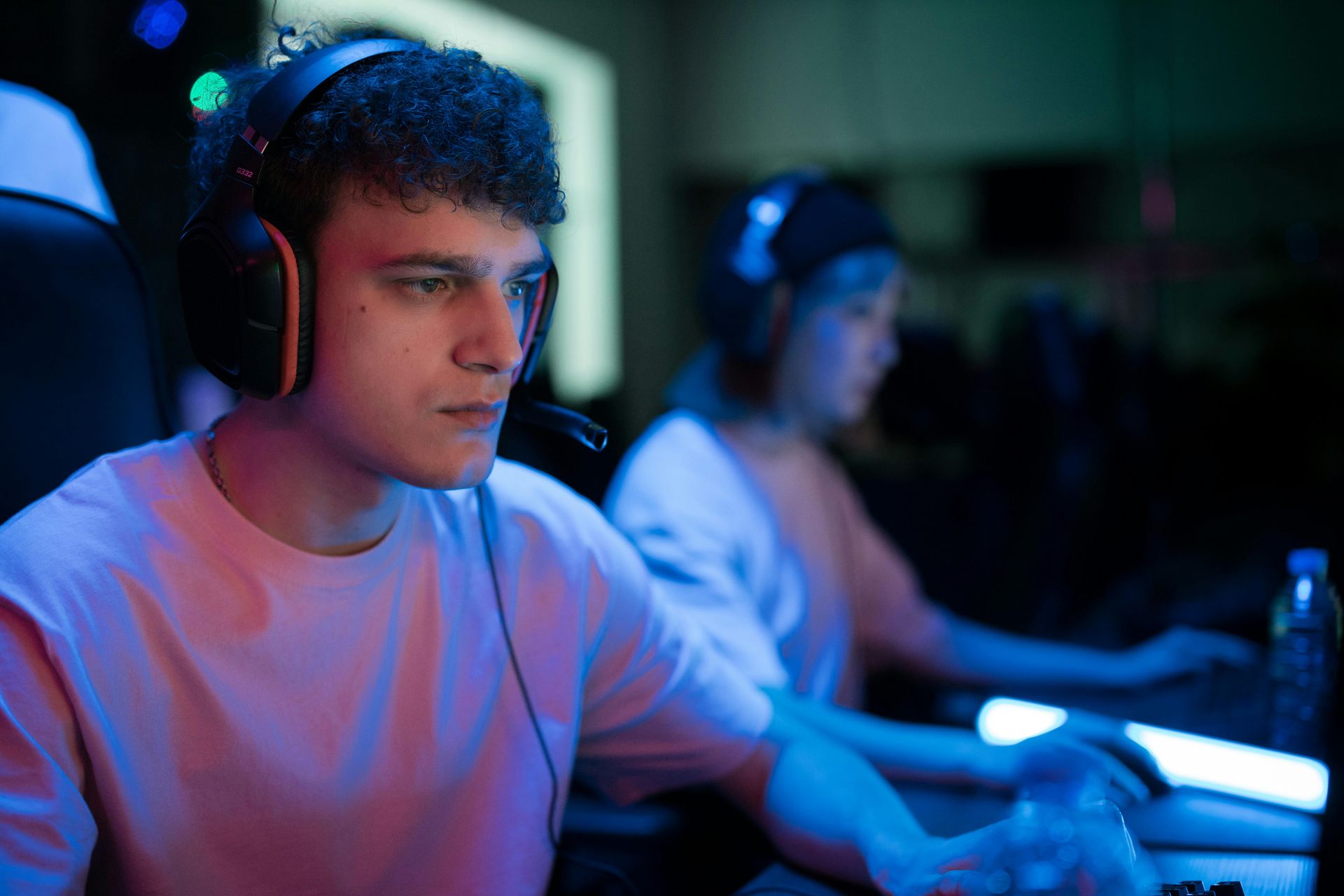 Man wearing headset at a computer, focused on the screen. Another person in background. Low-lit room, blue and pink lights.