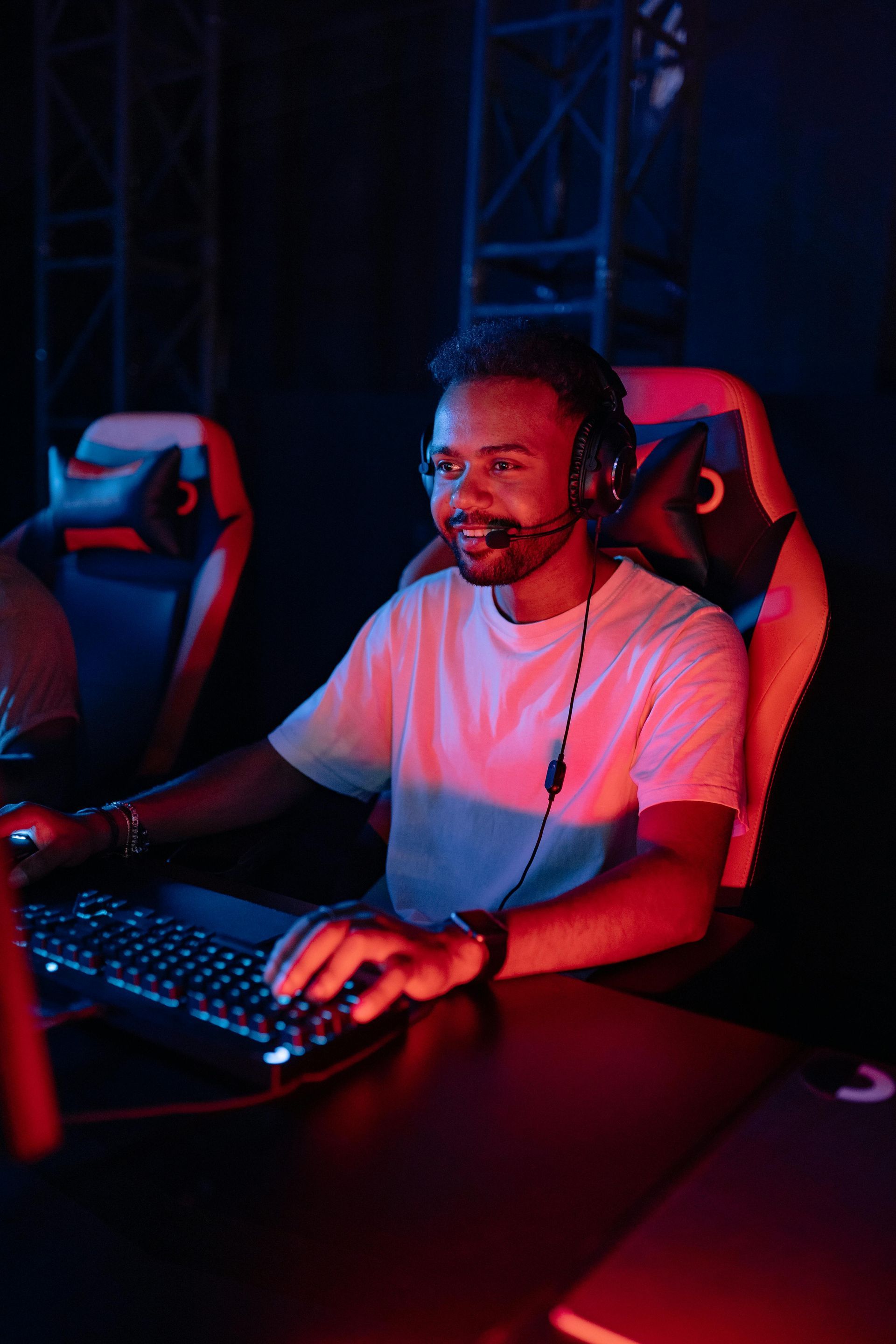 Man wearing a headset, smiling, playing at a computer with a glowing keyboard, in a dimly lit room with red accents.