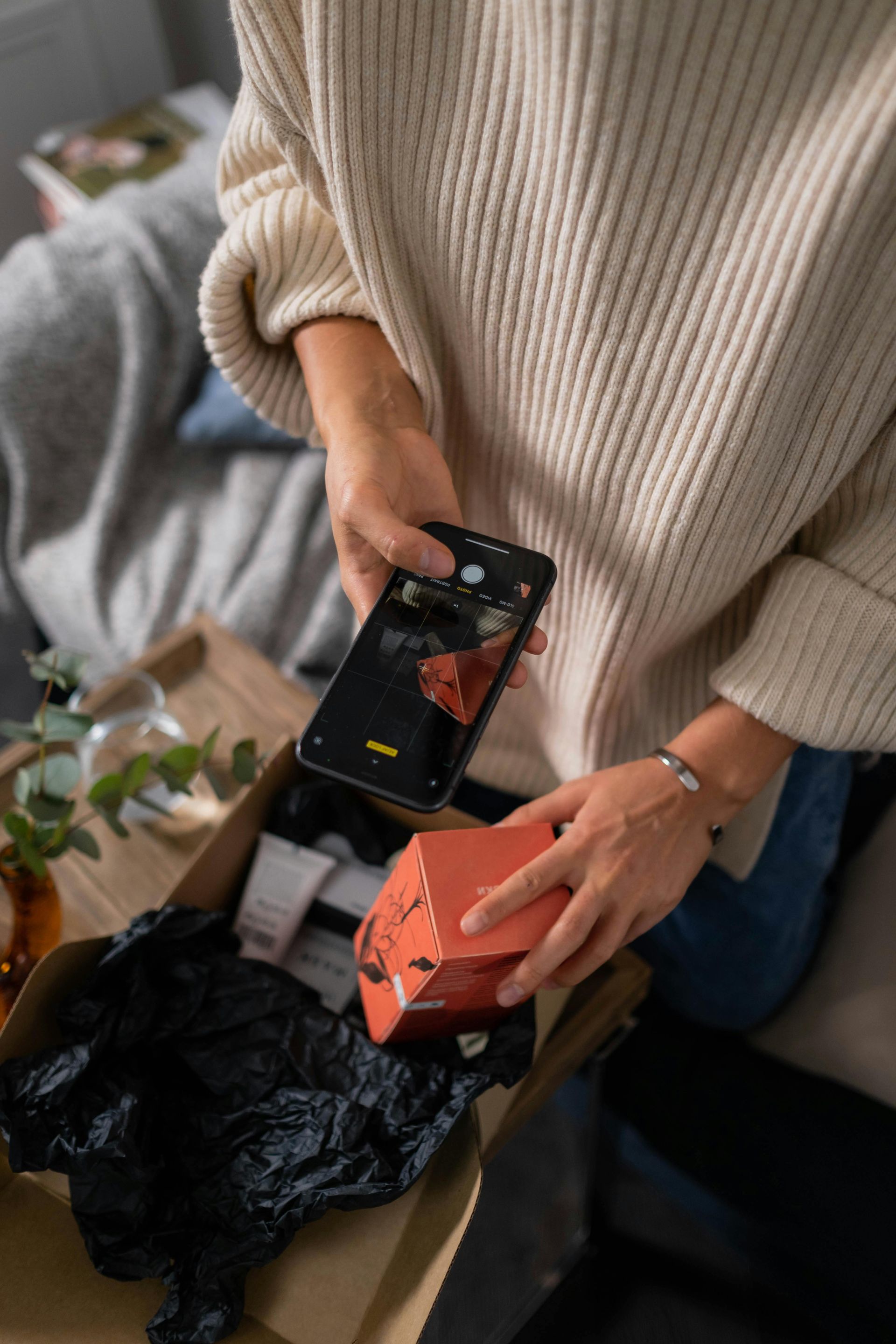 Person holding a phone over an orange box in a cardboard box with black tissue paper.