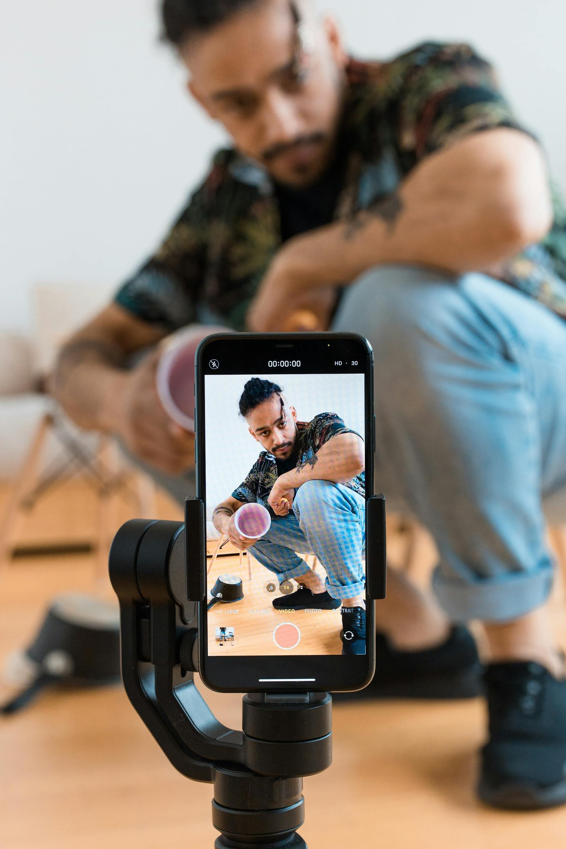 Man filming with a phone on a tripod, holding a container, squatting indoors.
