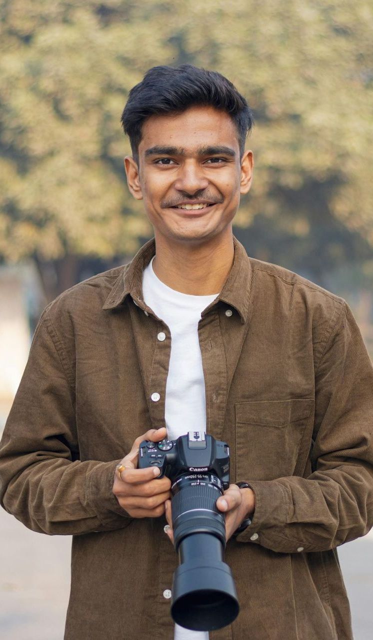 Man holding a camera, smiling. Wearing a brown corduroy shirt over a white shirt, outdoors.