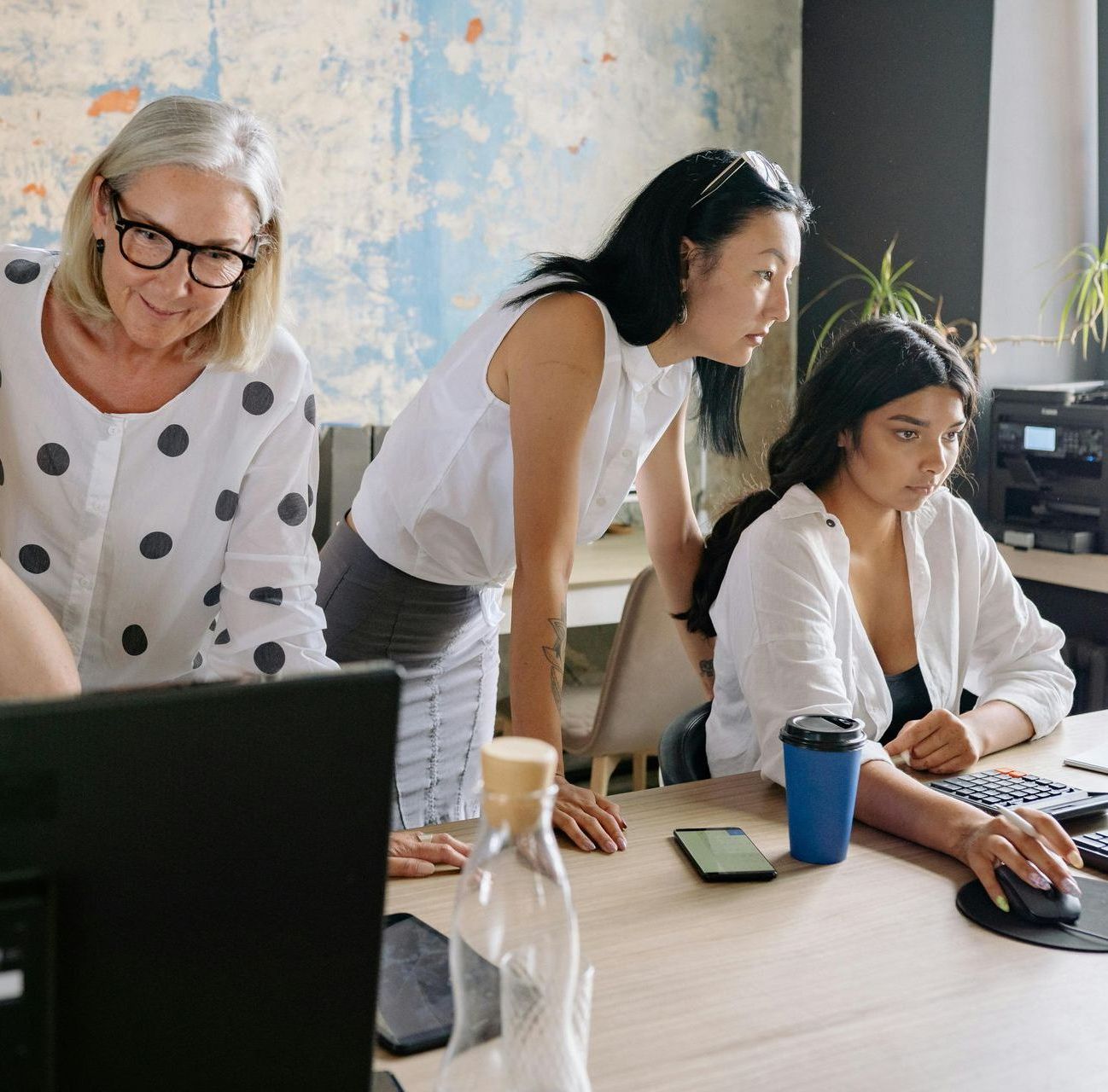 Three women at a desk looking at a computer monitor. One woman uses the mouse; two others observe.