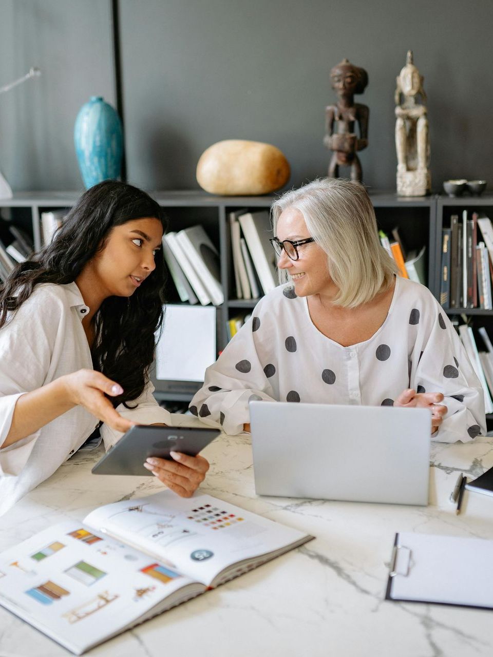 Woman showing a tablet to another woman, working at a table with a laptop, book, and clipboard in front of a bookshelf.