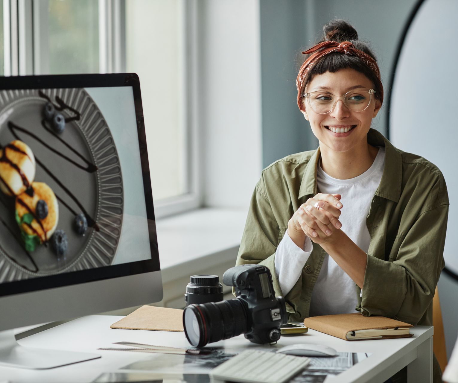Photographer with glasses smiles at the camera, a food photo on her computer, camera, and lens on the desk.