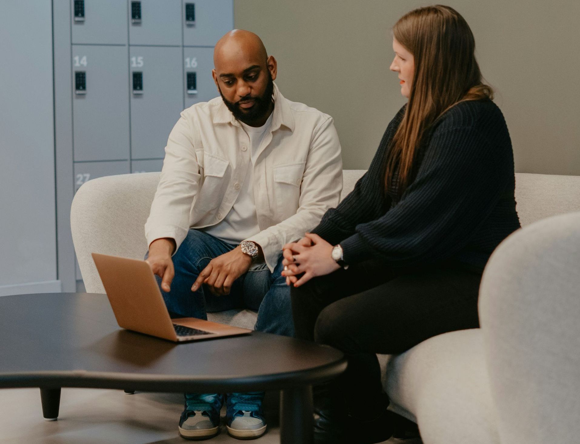 Man points at laptop screen, discussing with woman on a couch in a modern office.