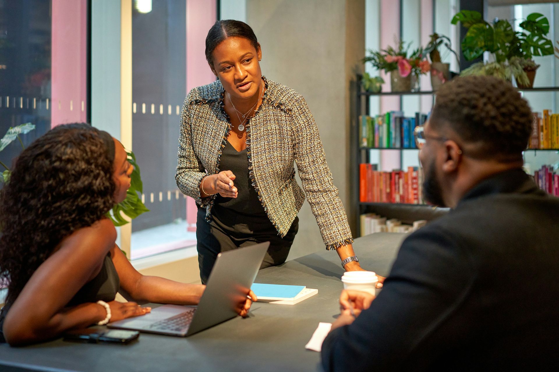 Three people in an office meeting: woman speaking, laptop open, bookshelf background.