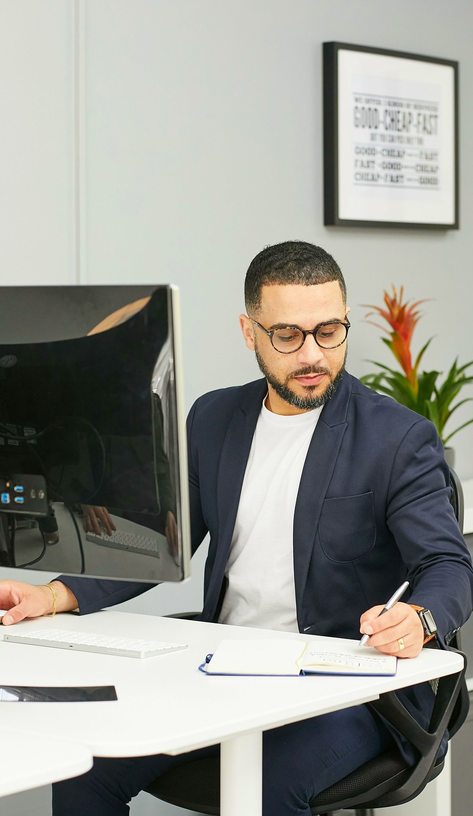 Man in glasses and blazer at desk, writing in notebook. Computer monitor, plant, and framed art in the background.