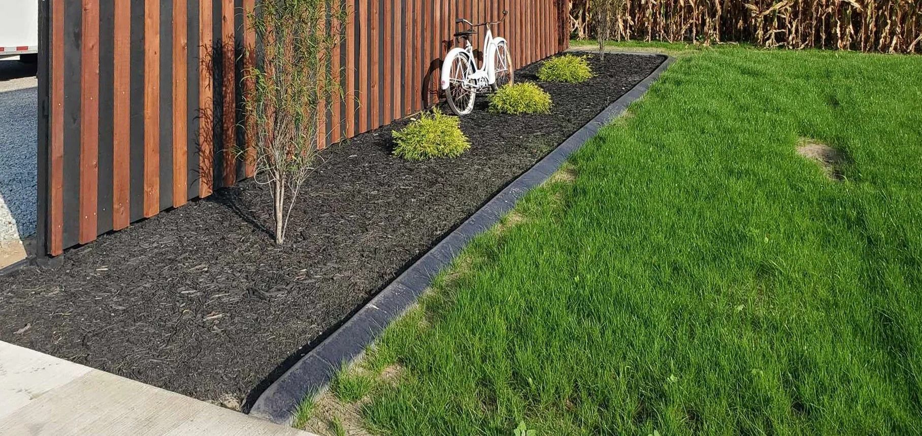 A black mulch bed with a black border separates green grass from a wooden fence and a concrete sidewalk.