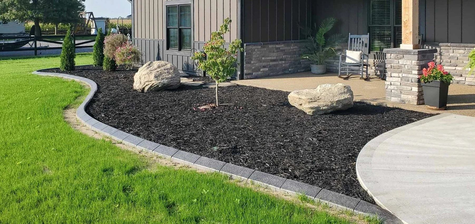 A yard with green grass, black mulch, gray border, and a home with a porch.