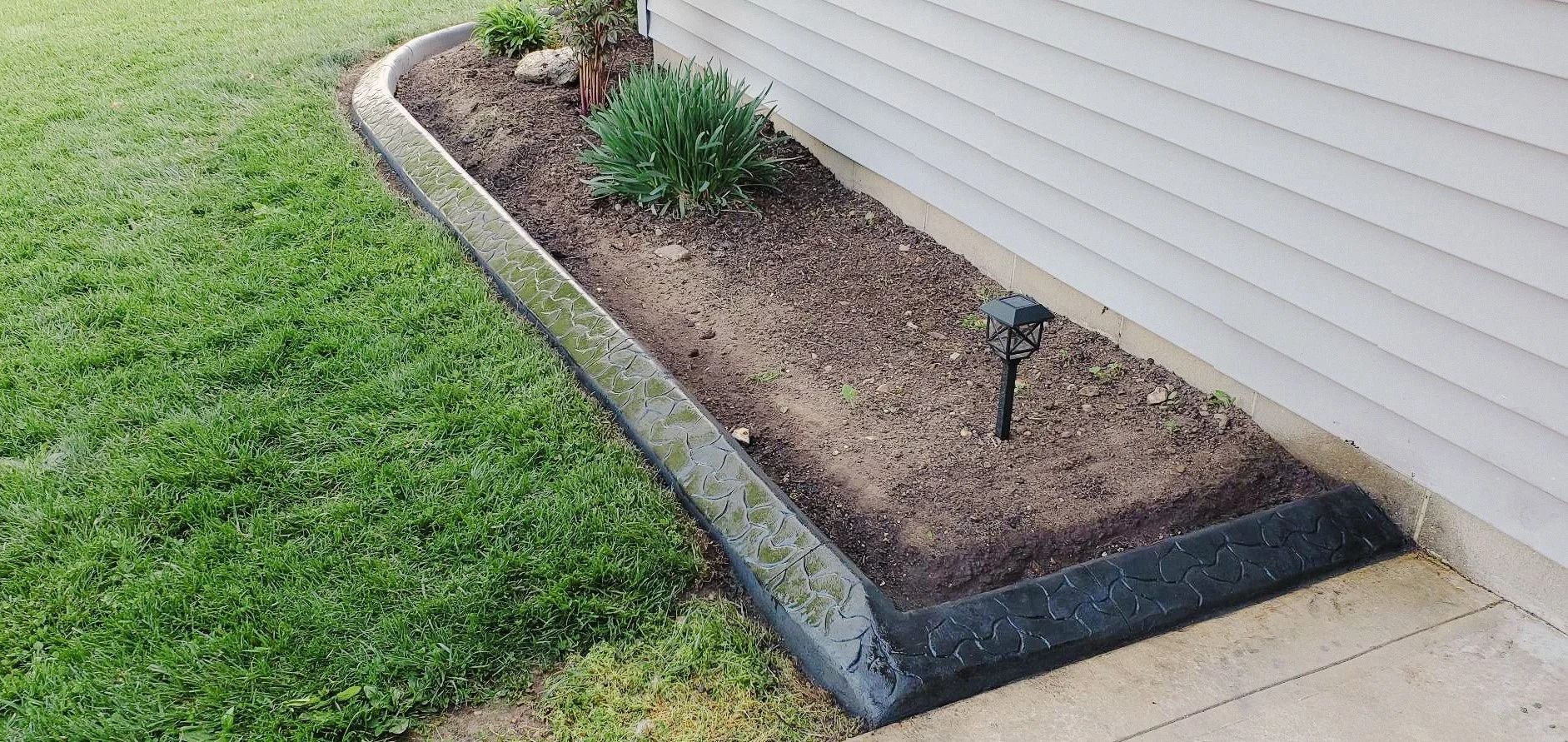 A rectangular flower bed with dark edging next to a building and lawn. The bed is filled with dark soil and plants.