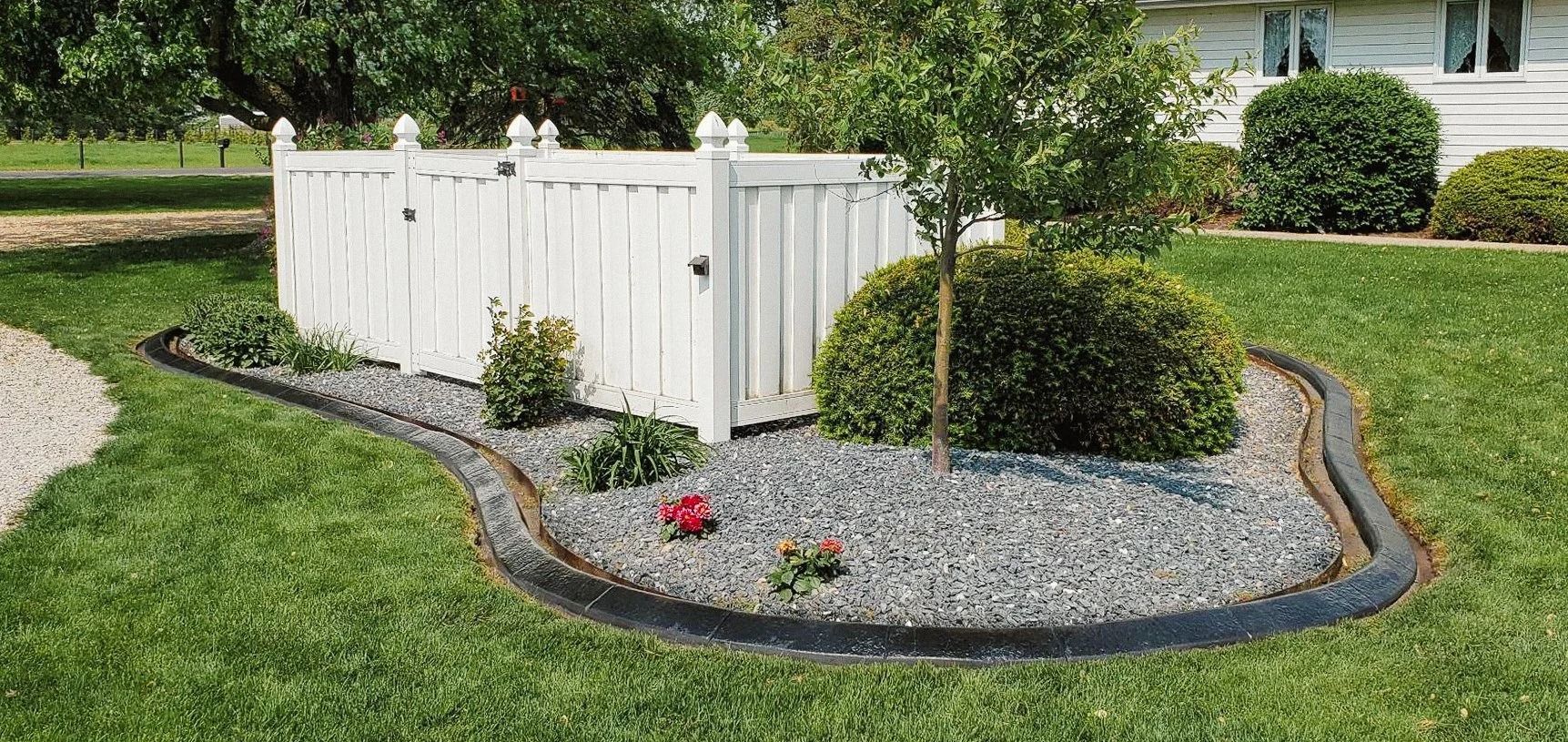 A white fence surrounds a landscaped area with dark edging, plants, and gray gravel on green grass.