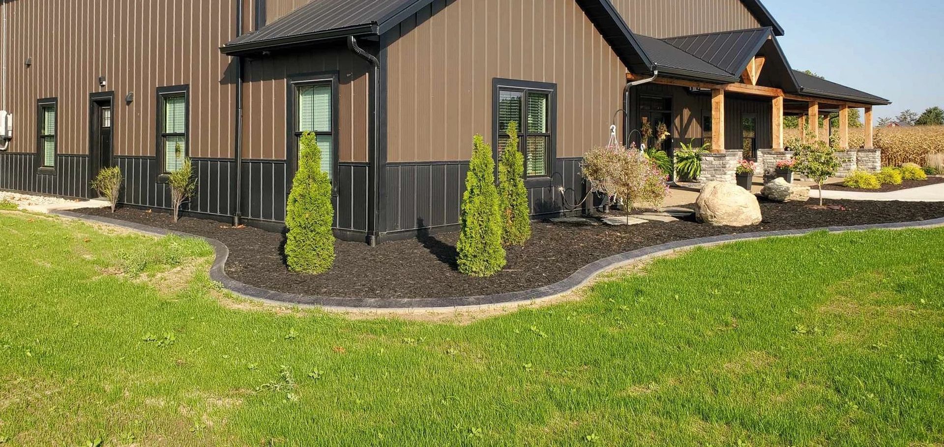 Brown building with black trim, a porch, green lawn, and dark mulch beds with small evergreen trees.