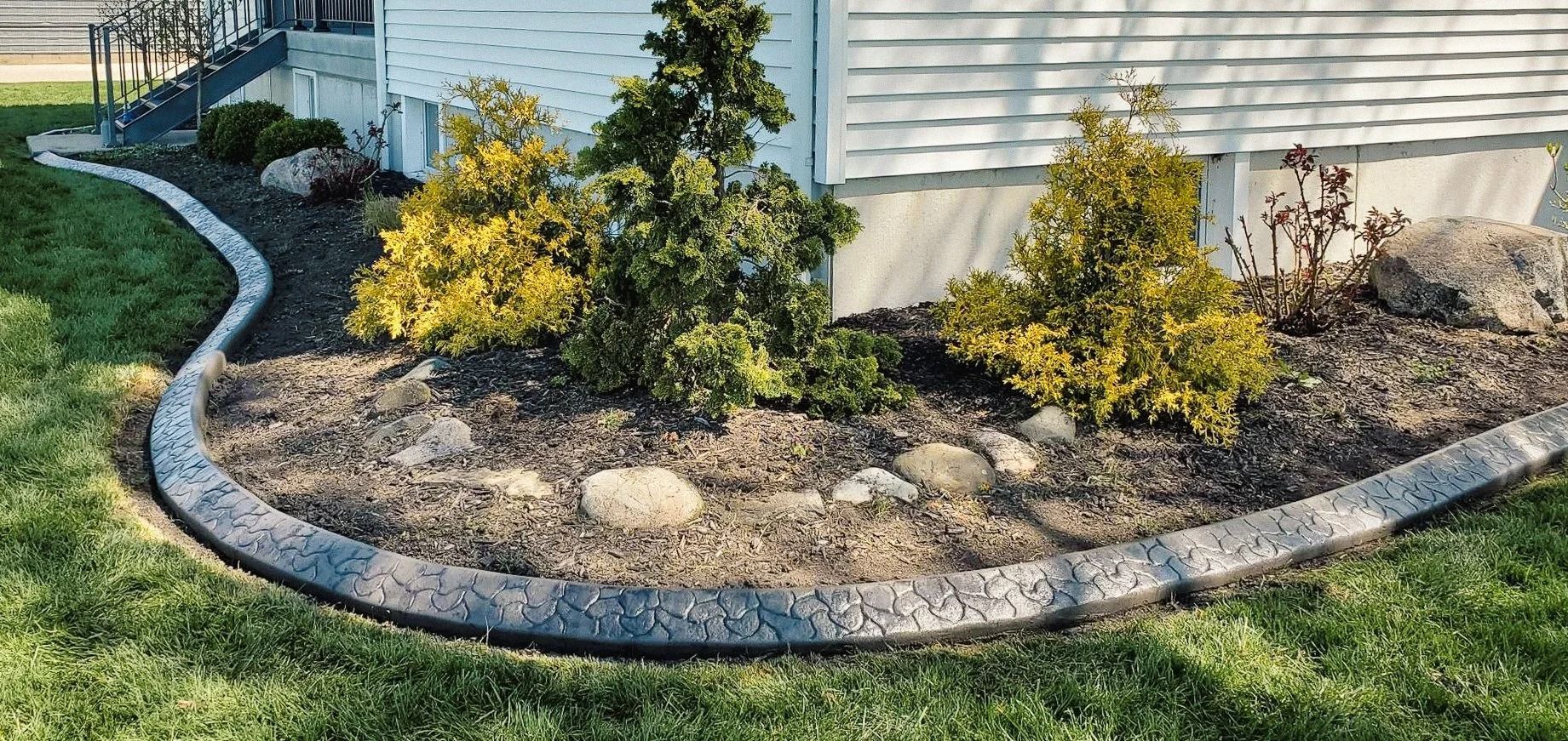 Curved concrete border edging a landscaped garden bed with shrubs and a house in the background.
