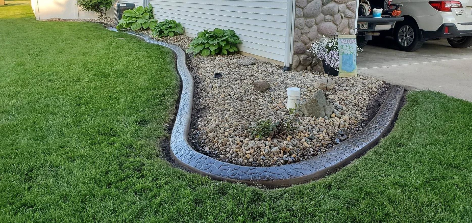 A landscaped yard with a concrete curb, gravel, and grass. A car is parked in the background.