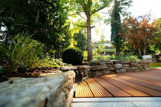 Stone wall and wooden deck in a sunlit garden with trees and shrubs.