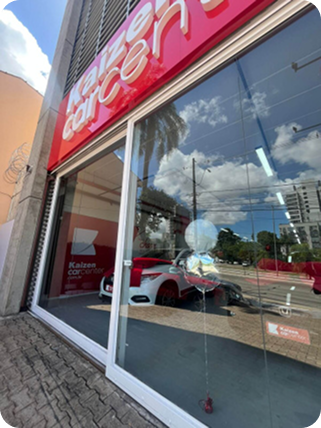 Fachada da concessionária de carros Kaizen, carro branco em exposição, com sinalização vermelha e branca.
