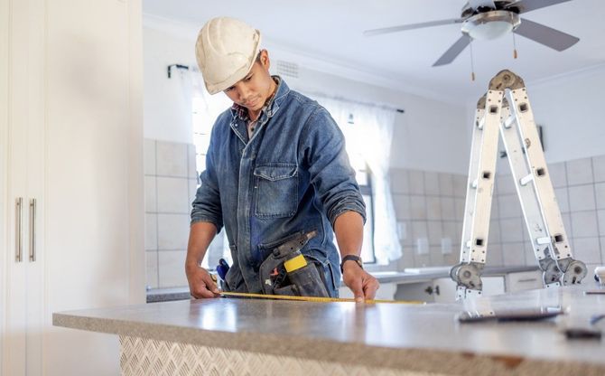 Worker in a hard hat measuring a countertop in a bright room, with a ladder and ceiling fan nearby.
