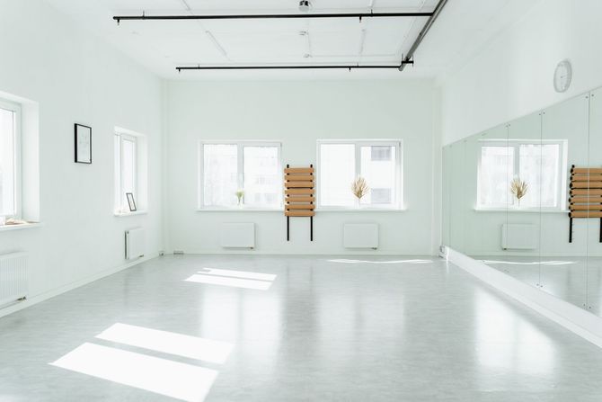 Bright white dance studio with mirrors, windows, barre, and a wooden chair centered on the back wall.