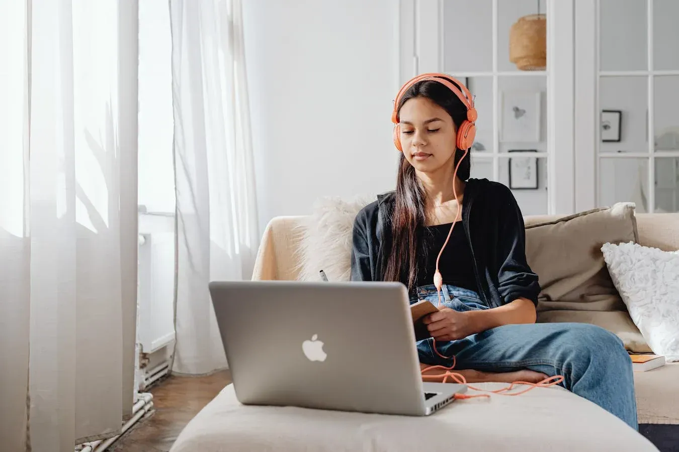 Person wearing headphones uses a laptop while sitting on a couch. Bright, sunlit room.