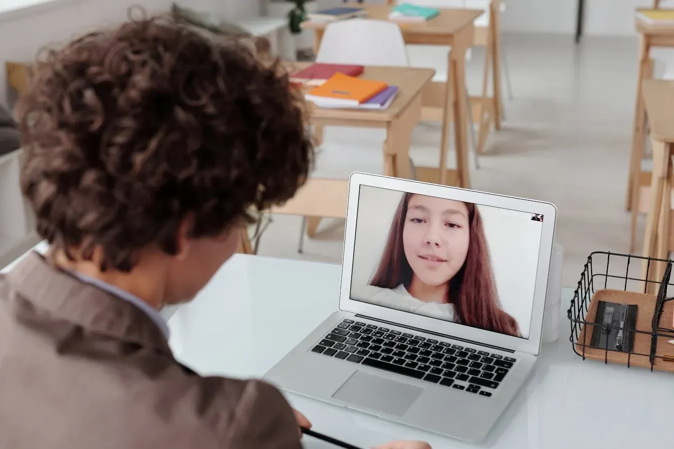 A person with curly hair views a video call on a laptop in a classroom. The screen shows another person smiling.