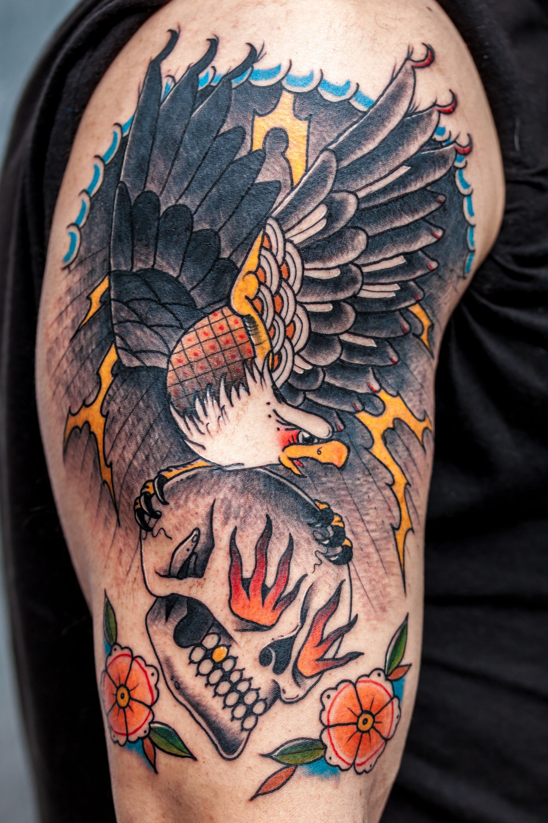 Tattoo of a black and orange eagle perched over a skull with red handprints and flowers on an arm