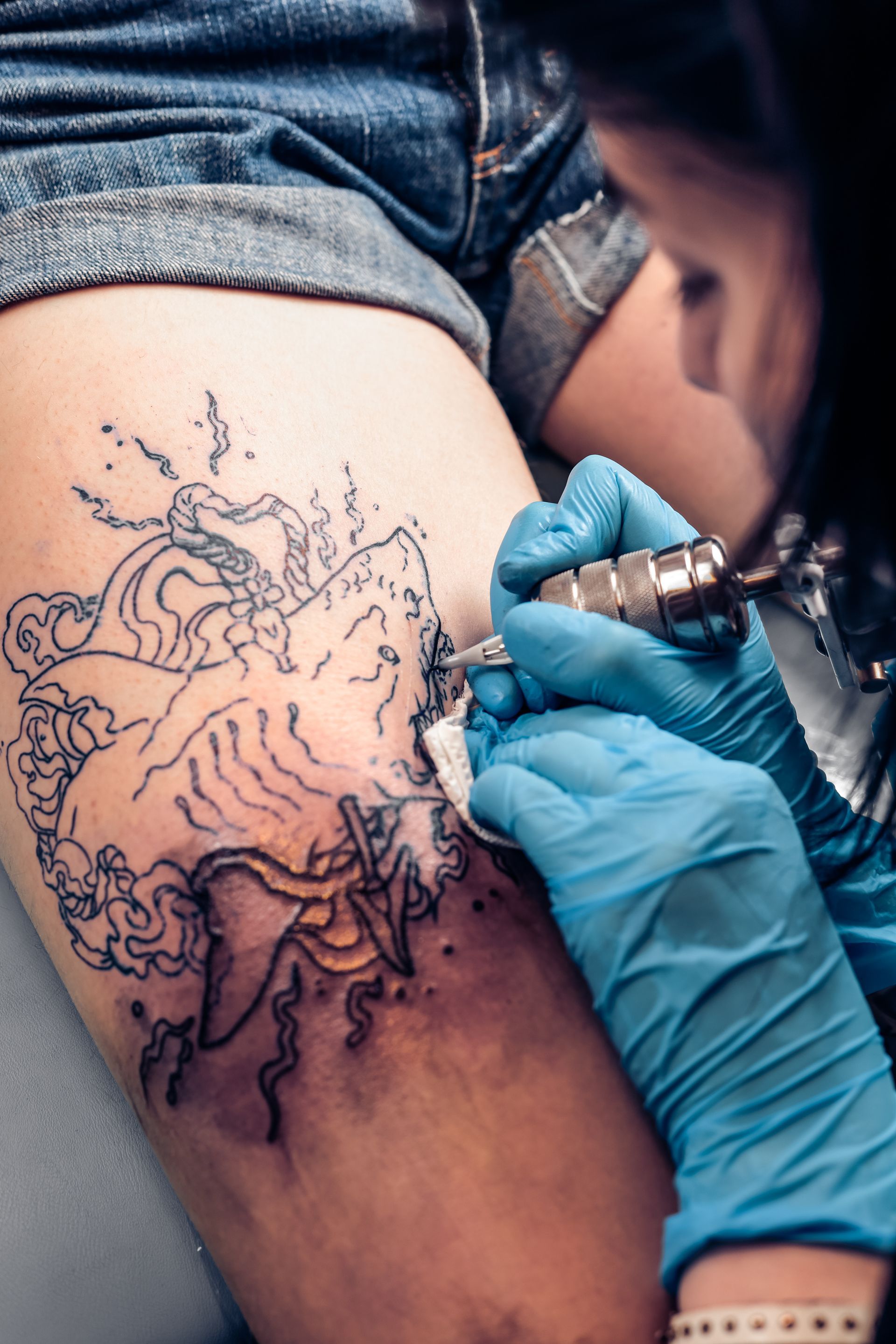 Artist outlining a black ink tattoo on a person’s upper thigh with blue-gloved hands
