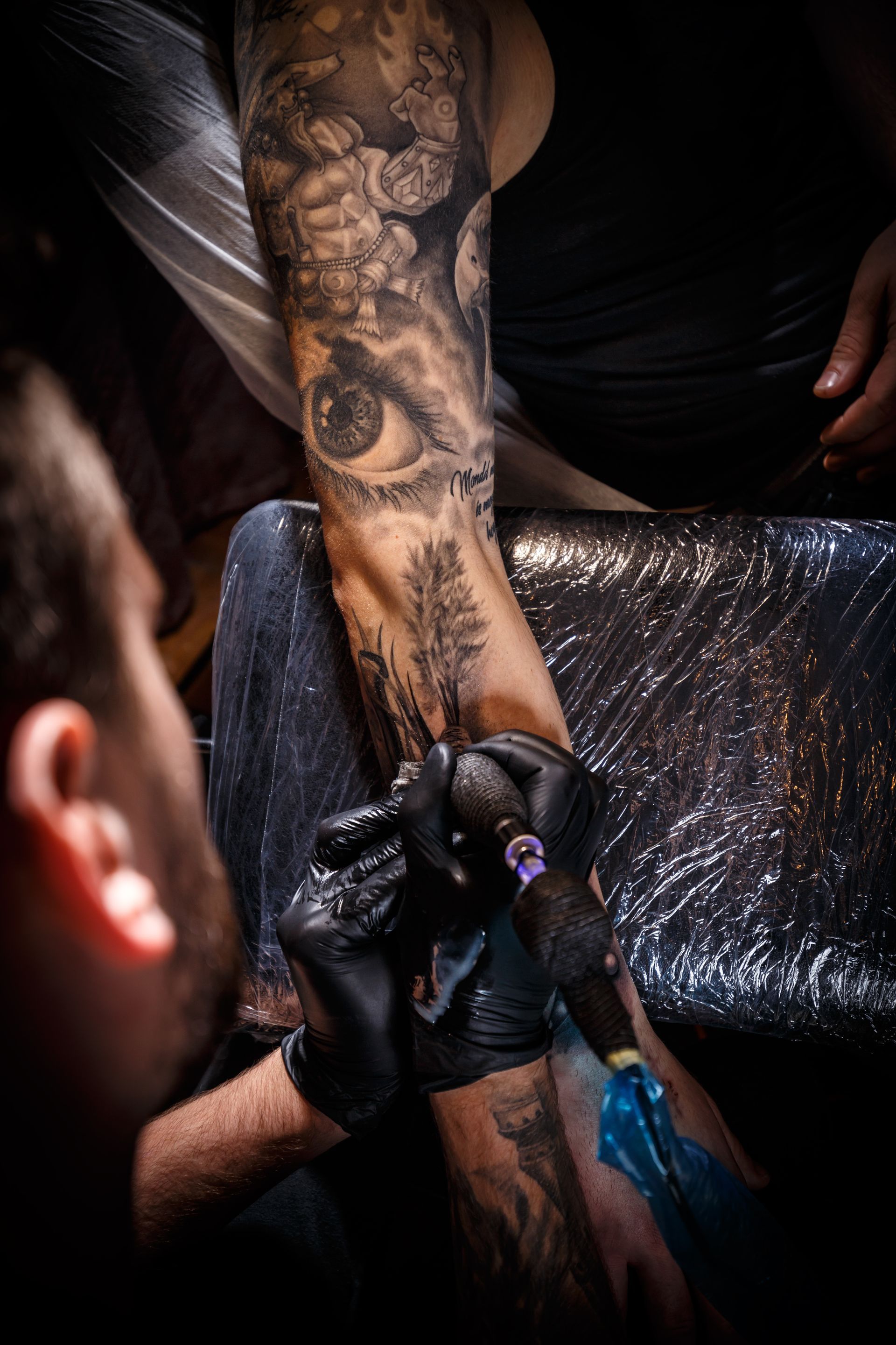 Tattooed forearm being inked with a tattoo machine by gloved artist in a dark studio