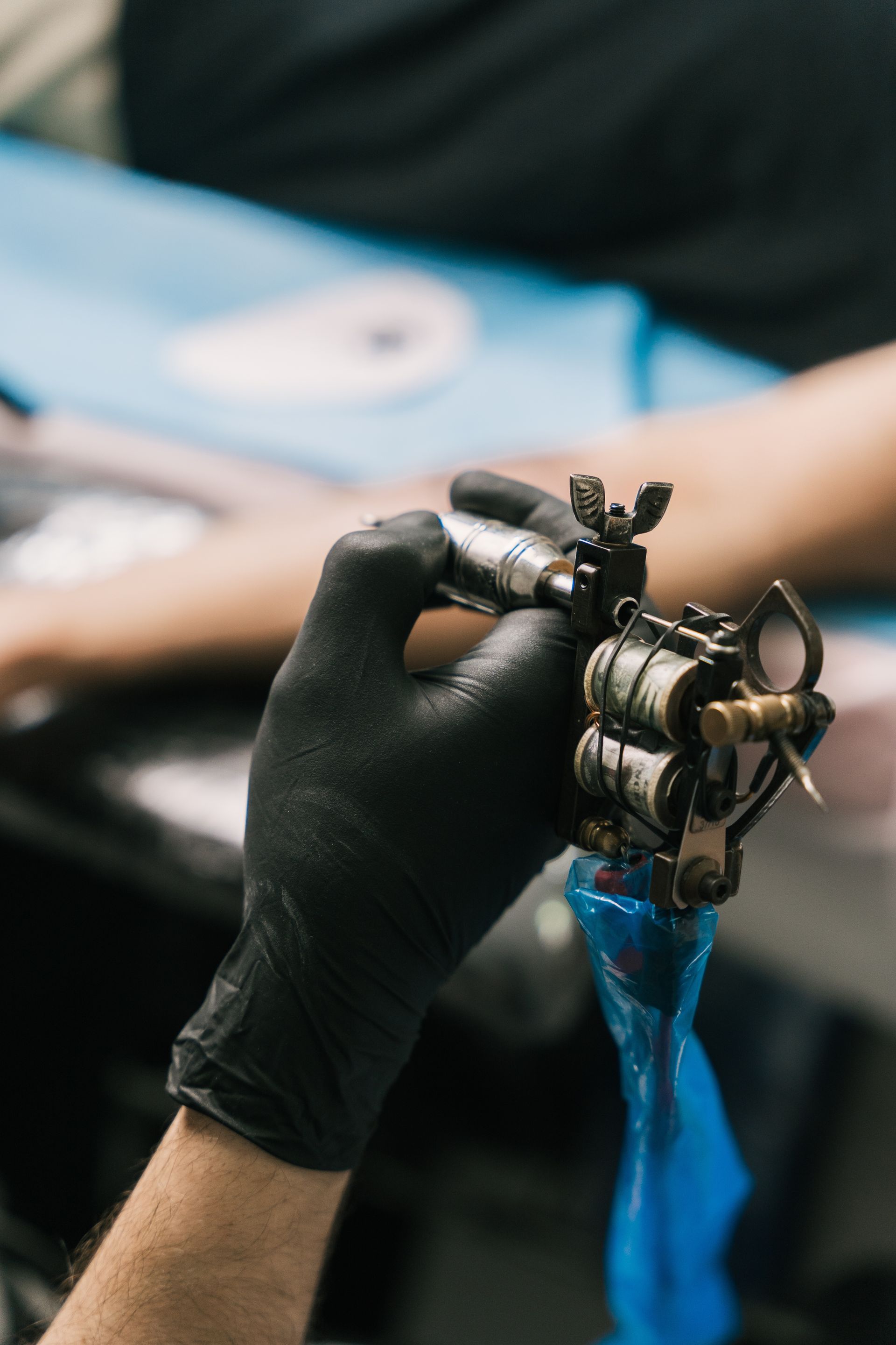 Black-gloved hand holding a tattoo machine with blue ink dripping from the needle