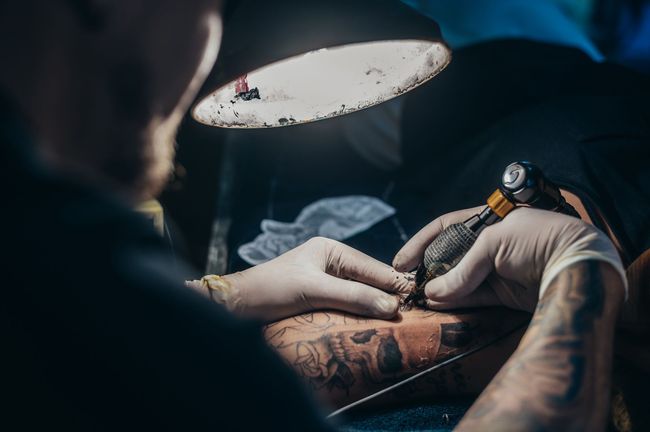 Tattoo artist in gloves tattooing a client’s forearm under a bright lamp