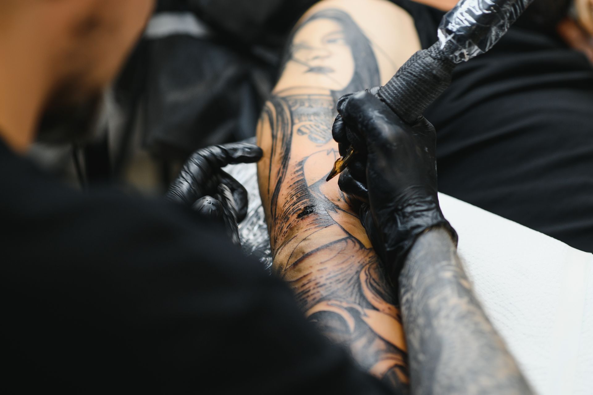 Tattoo artist working on a black-and-gray tattoo on a client’s arm, close-up with gloves and ink pen